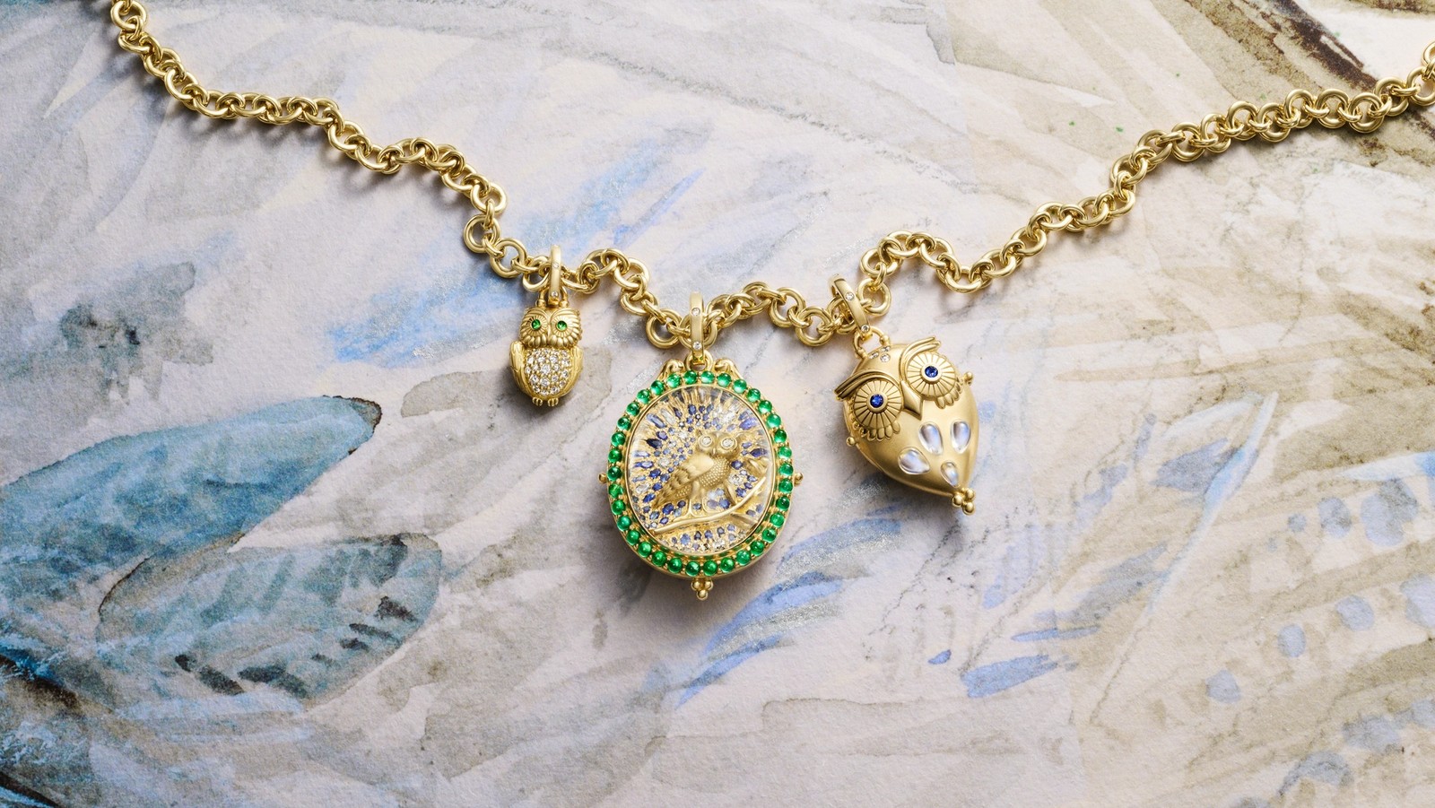 A gold necklace with three charms: a small diamond-bellied owl with green eyes, a round green-bordered bird pendant with blue and white gems, and a larger gold owl with blue eyes.