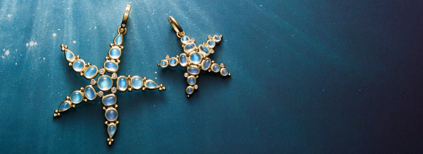 Two gold starfish pendants with opalescent blue moonstones and diamonds on a blue background with subtle light rays.