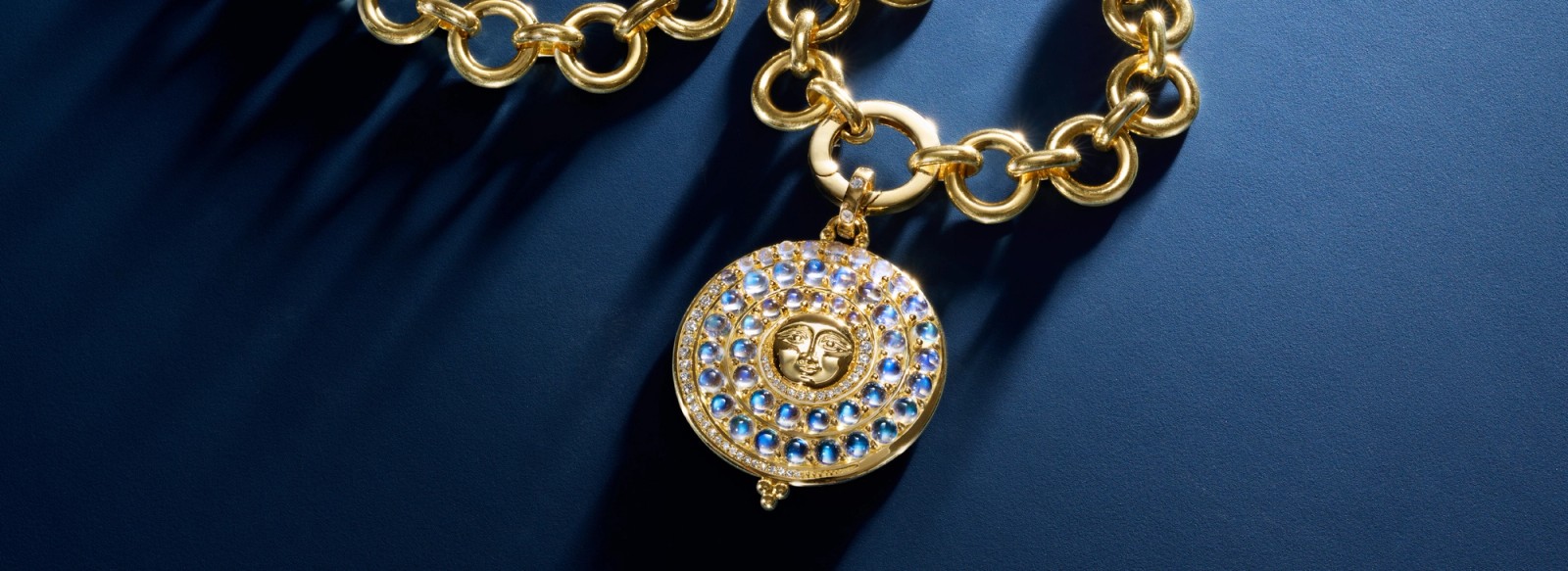Gold chain necklace with an ornate circular pendant featuring blue gems, diamonds, and a sun face on a dark blue background.