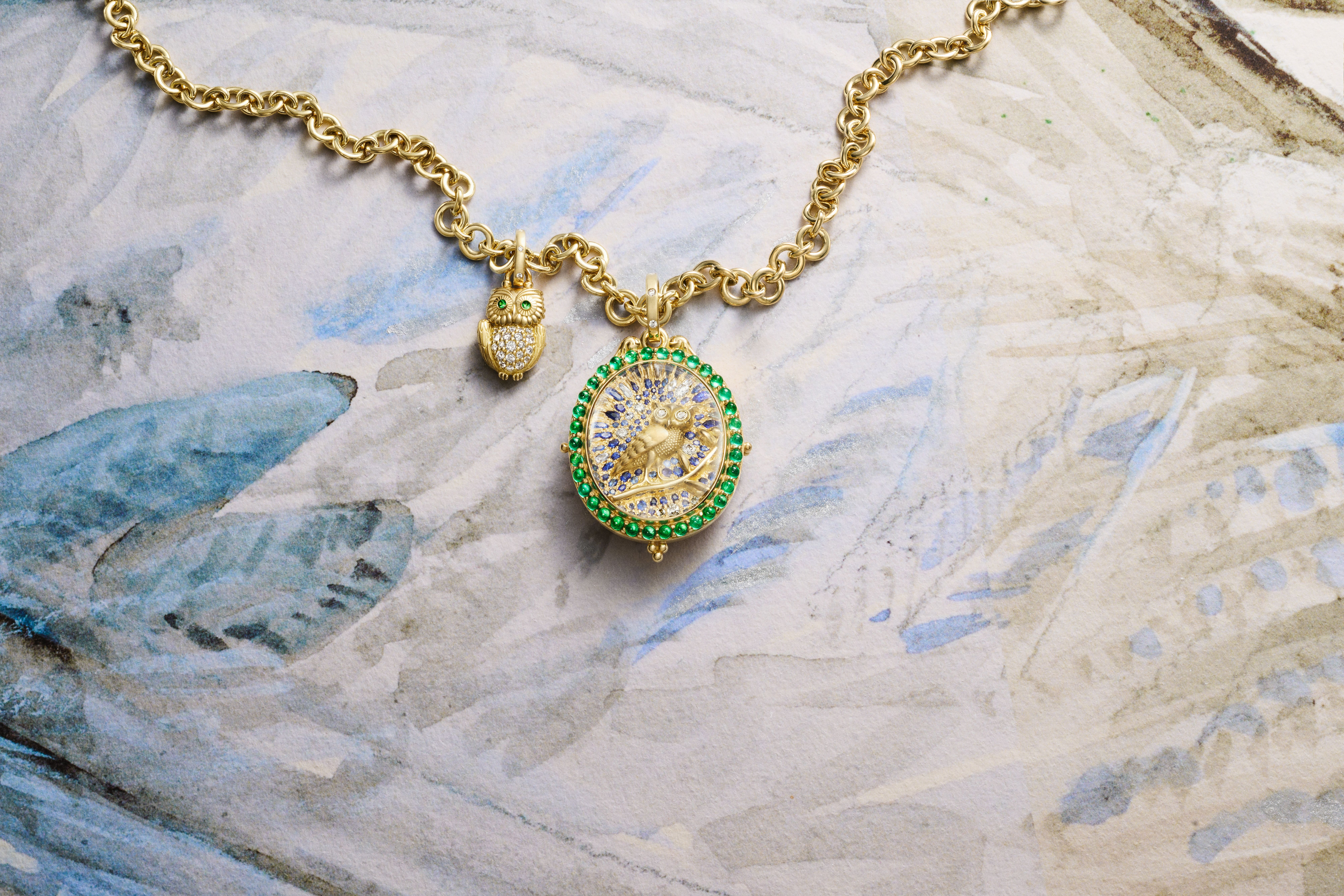Gold necklace with a pave diamond owl charm and an emerald-bordered locket featuring a bee, on a watercolor surface.