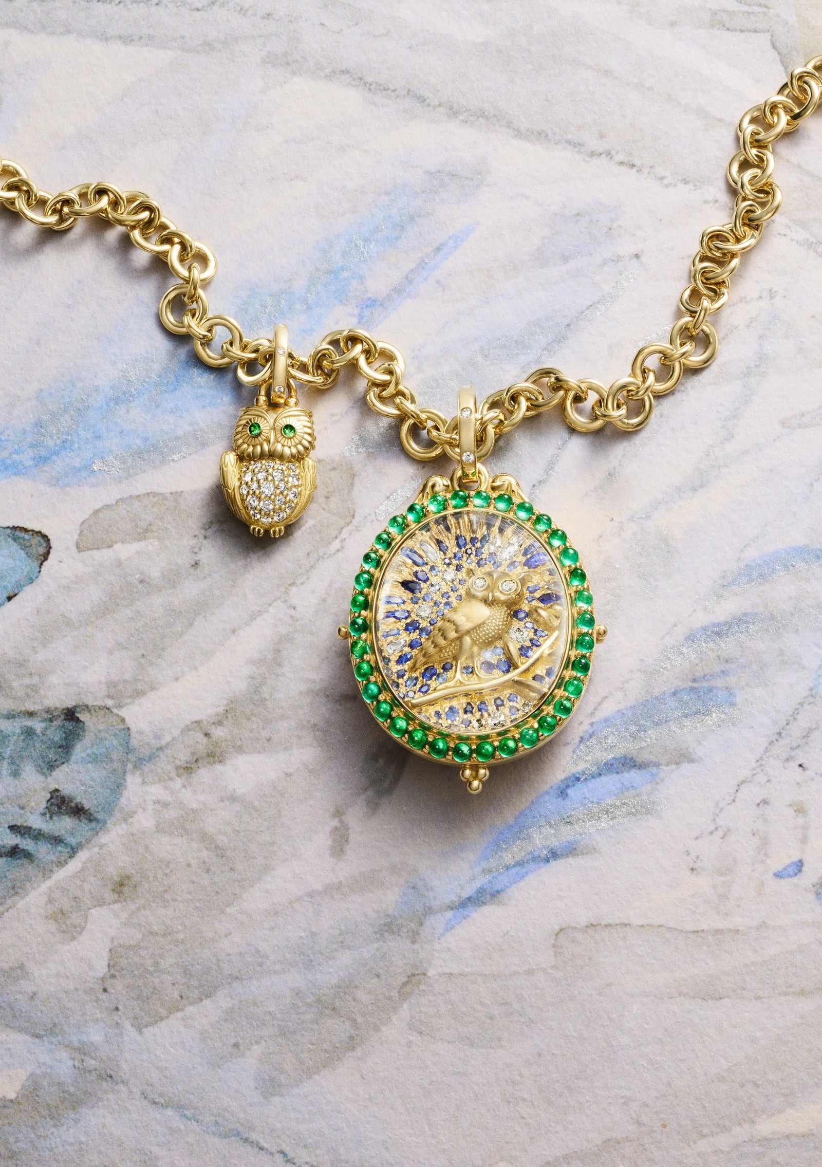 Gold necklace with a pave diamond owl charm and an emerald-bordered locket featuring a bee, on a watercolor surface.