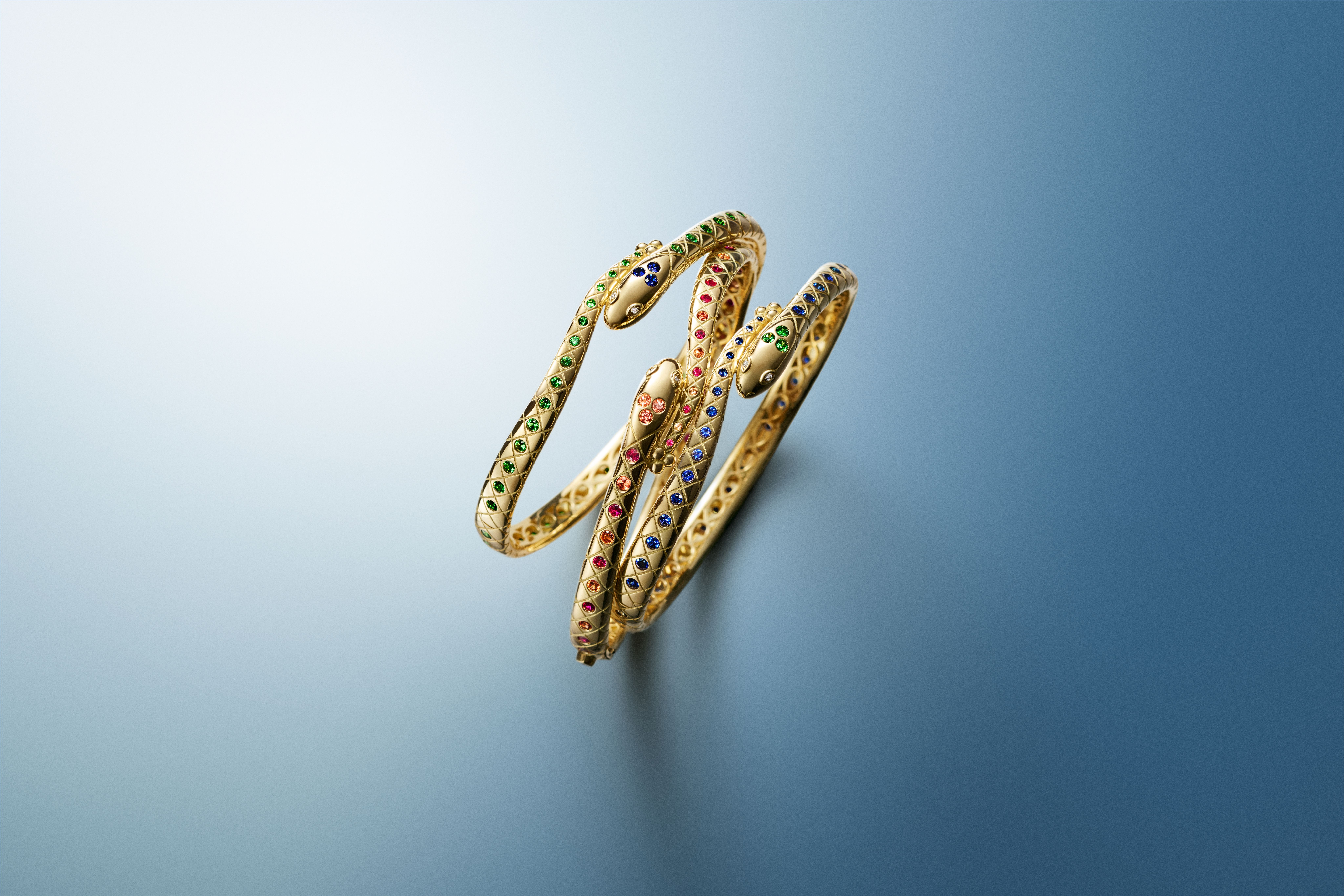 Three intertwined gold snake bangles, adorned with green, red, and blue gemstones, on a blue gradient background.