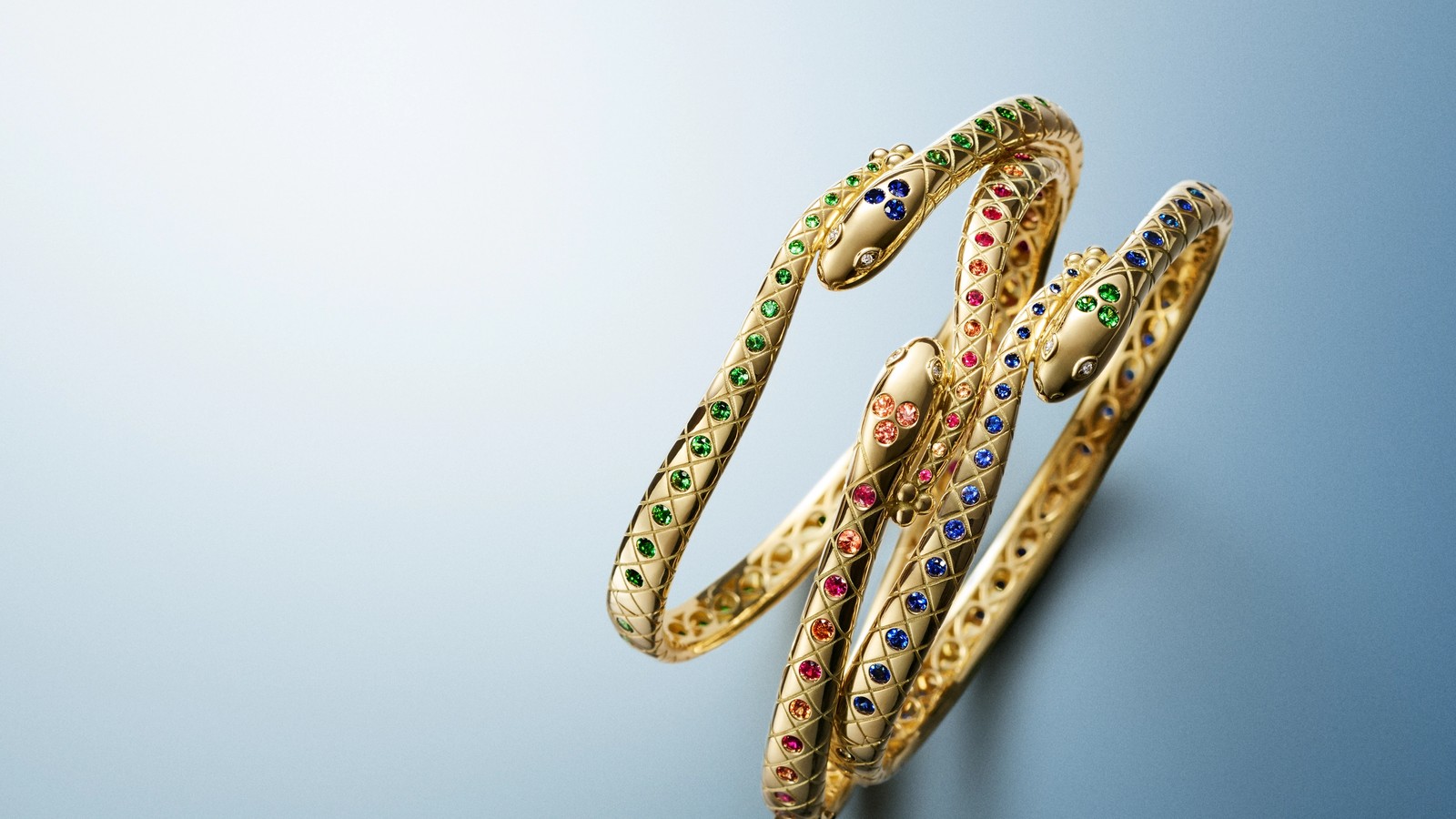 Three intertwined gold snake bangles, adorned with green, red, and blue gemstones, on a blue gradient background.