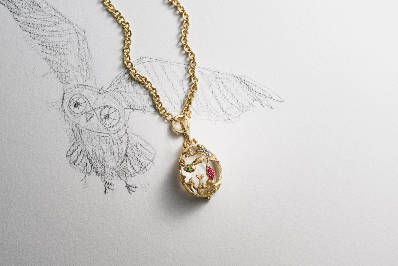 A gold chain necklace with an oval, jeweled nature-themed pendant lies over a pencil sketch of an owl.