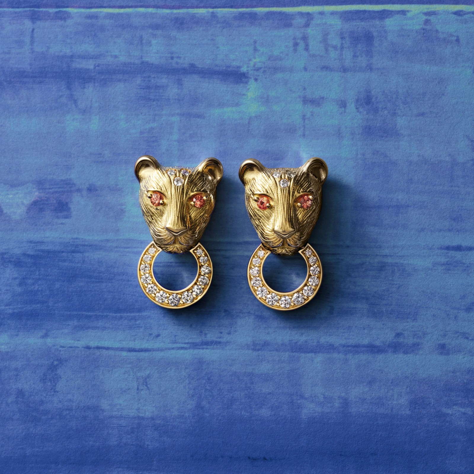 A pair of golden jaguar head earrings with pink gem eyes and diamond-studded C-rings on a blue background.