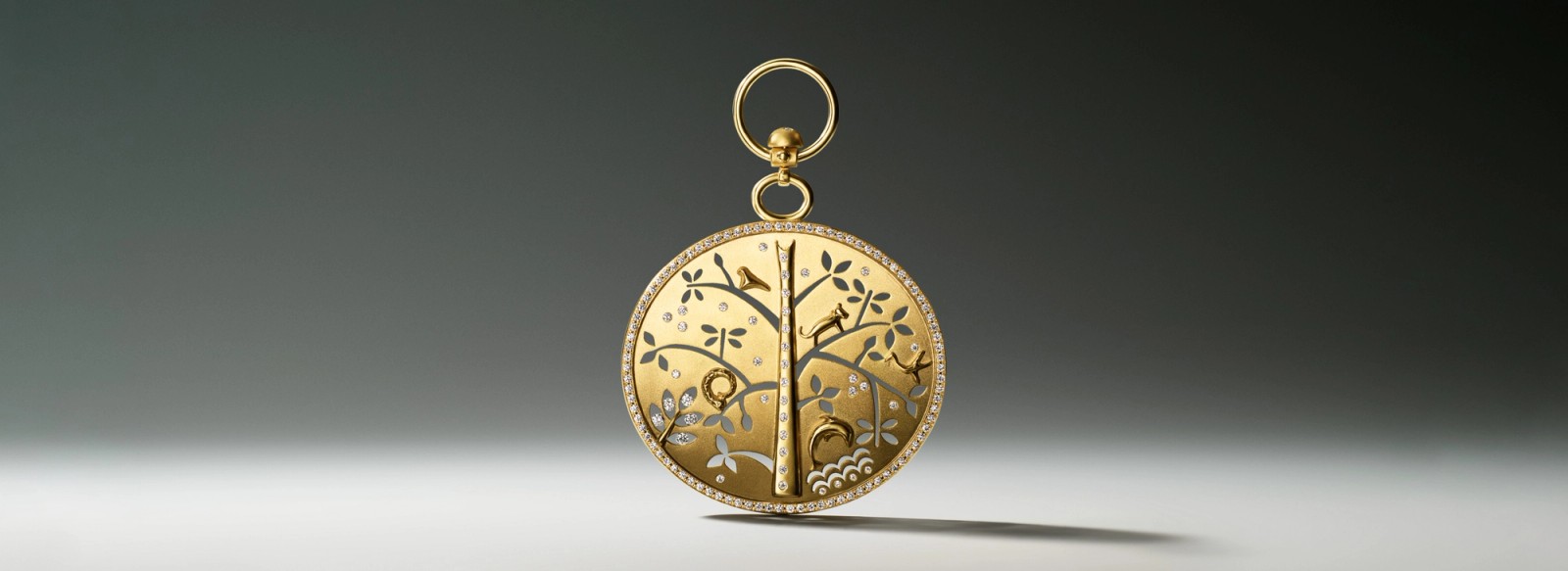 Gold round pendant featuring an ornate tree scene with birds, a cat, and silver leaves, set within a diamond border.