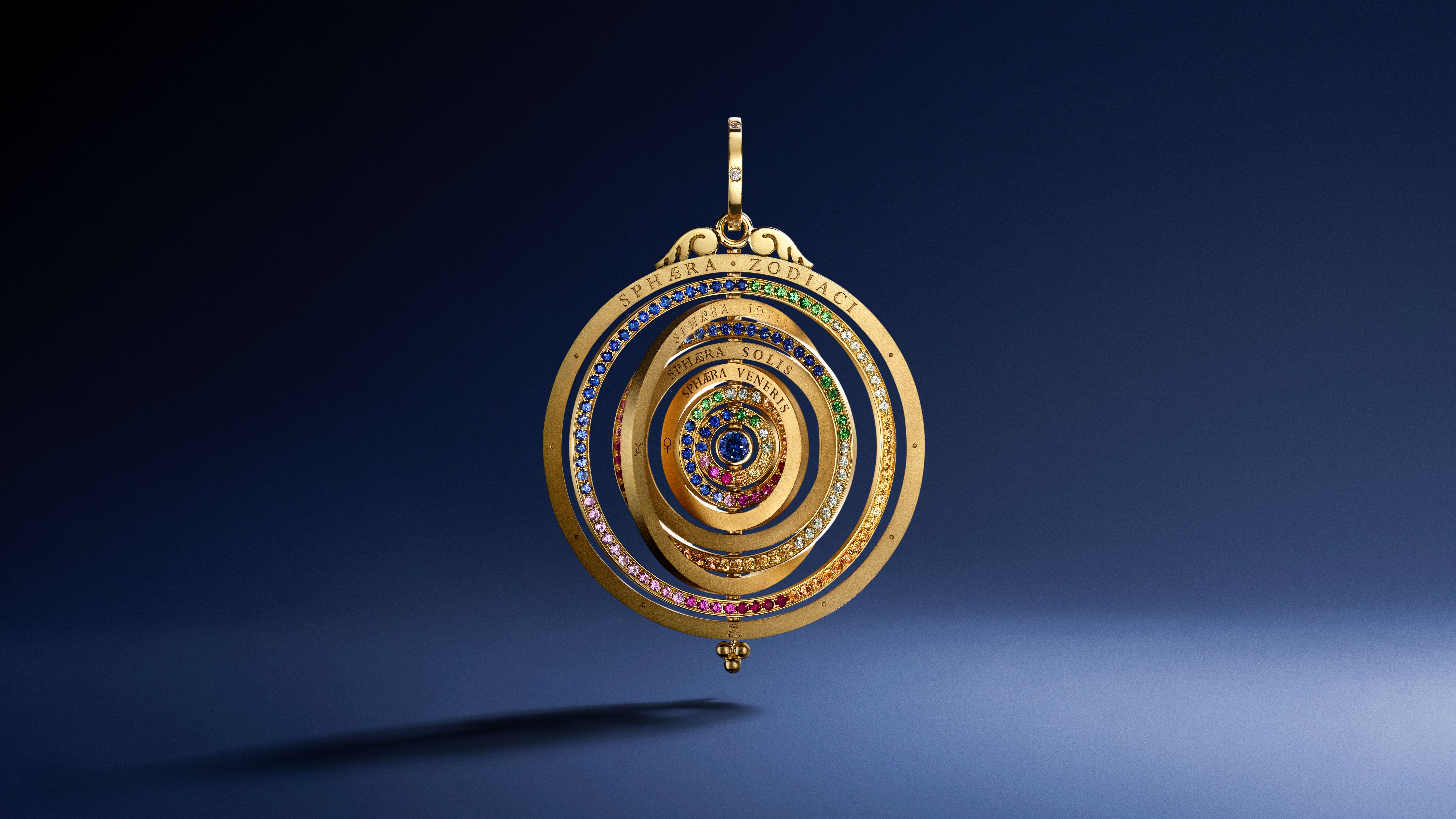 A gold pendant featuring concentric rings set with rainbow-colored gemstones, depicting a celestial model, on a dark blue background.