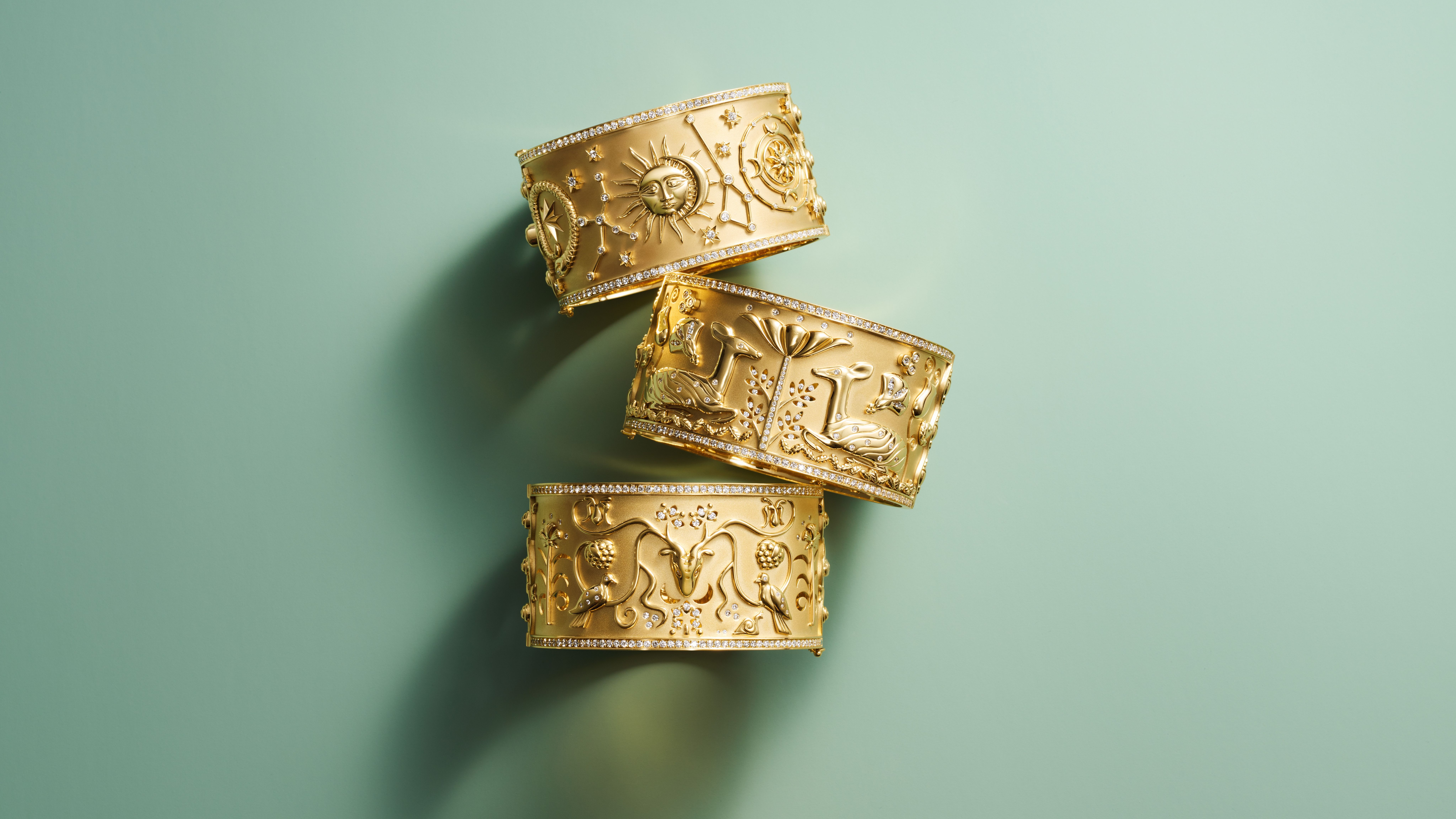 Three richly detailed gold cuffs with diamond edges are stacked on a teal background.