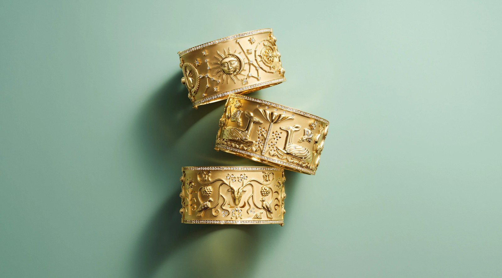 Three richly detailed gold cuffs with diamond edges are stacked on a teal background.