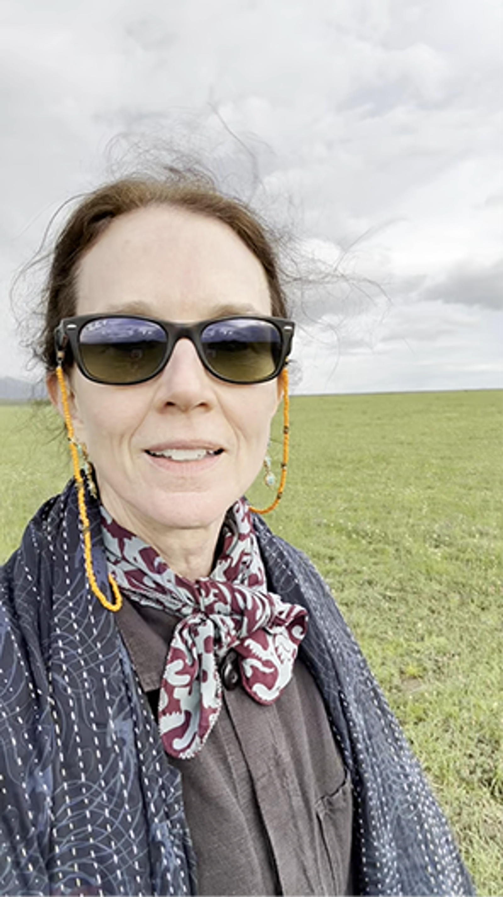 A woman with windswept hair, wearing sunglasses and a patterned scarf, takes a selfie in a green field under a cloudy sky.