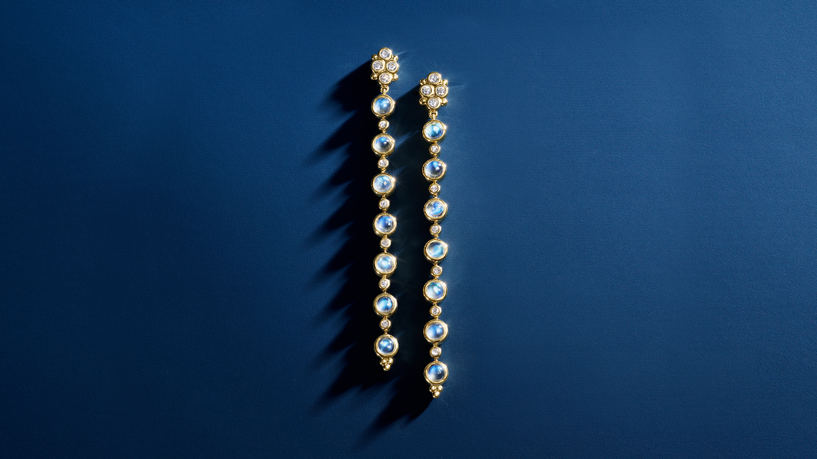 A pair of long gold dangle earrings with iridescent blue stones and diamonds on a dark blue background.