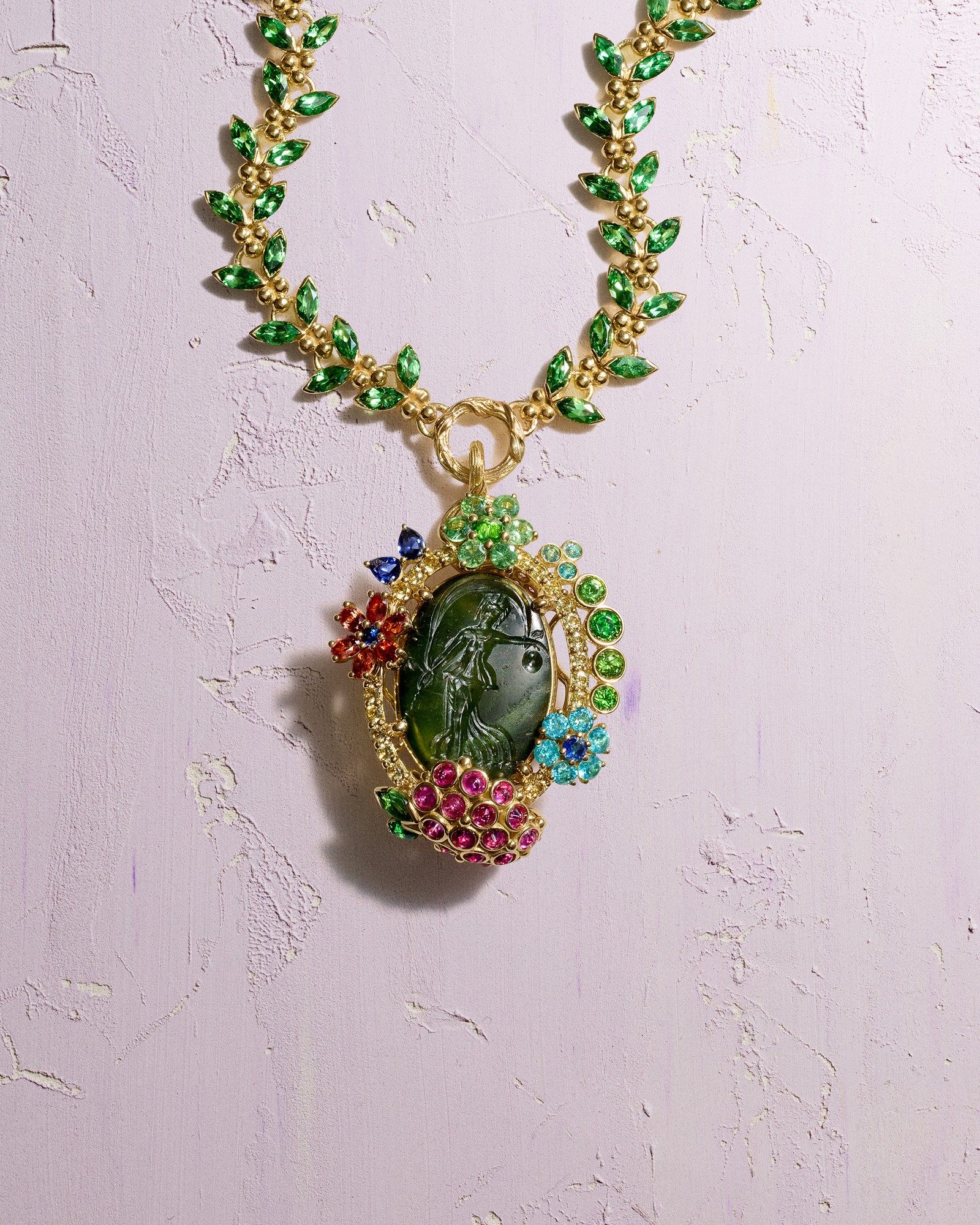 A gold necklace with green leaf-like gems and a pendant featuring a carved green cameo of a woman surrounded by multi-colored gemstone flowers, on a textured lavender surface.