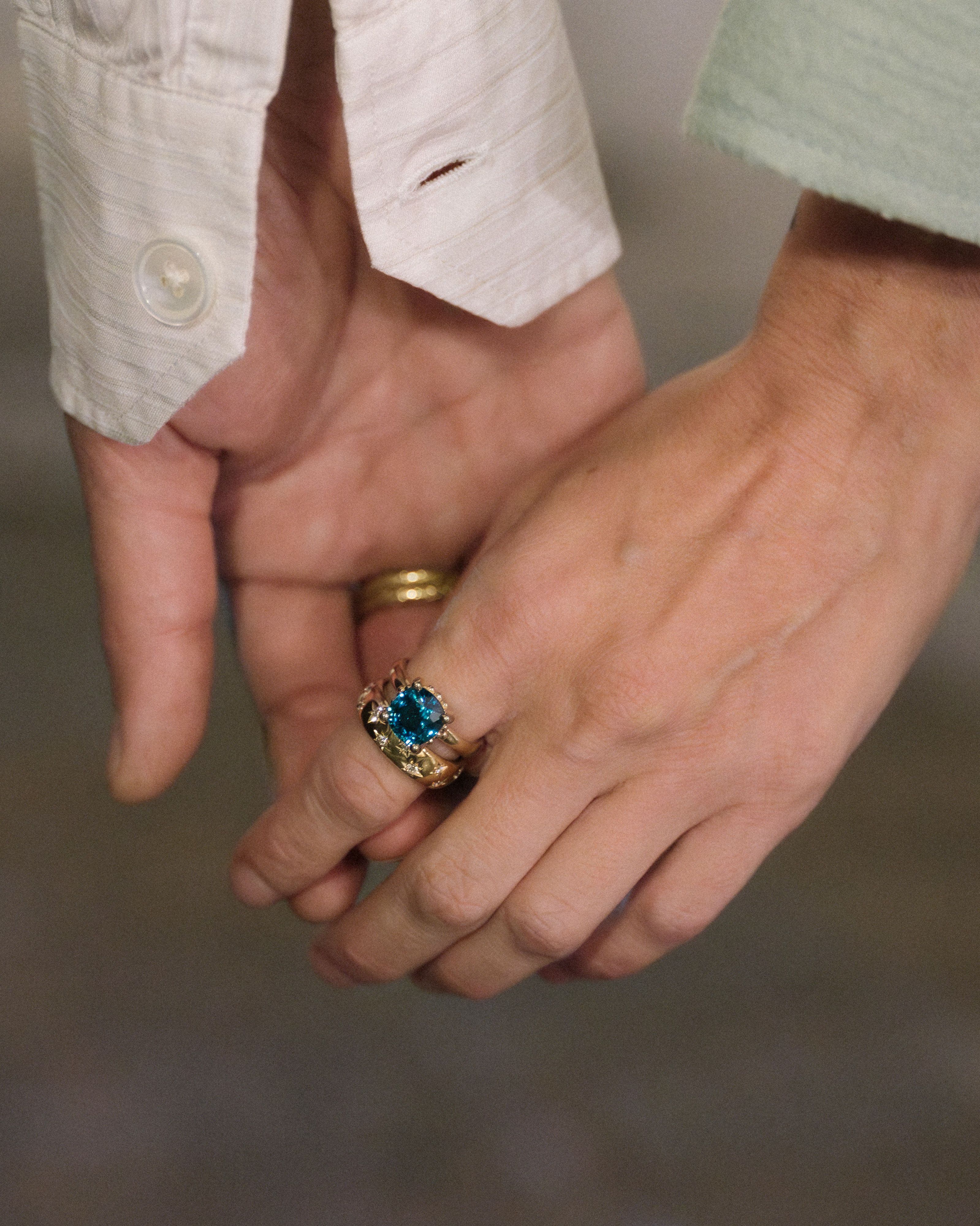 Two hands gently clasped, one wearing gold bands and the other a gold ring with a large blue gemstone.