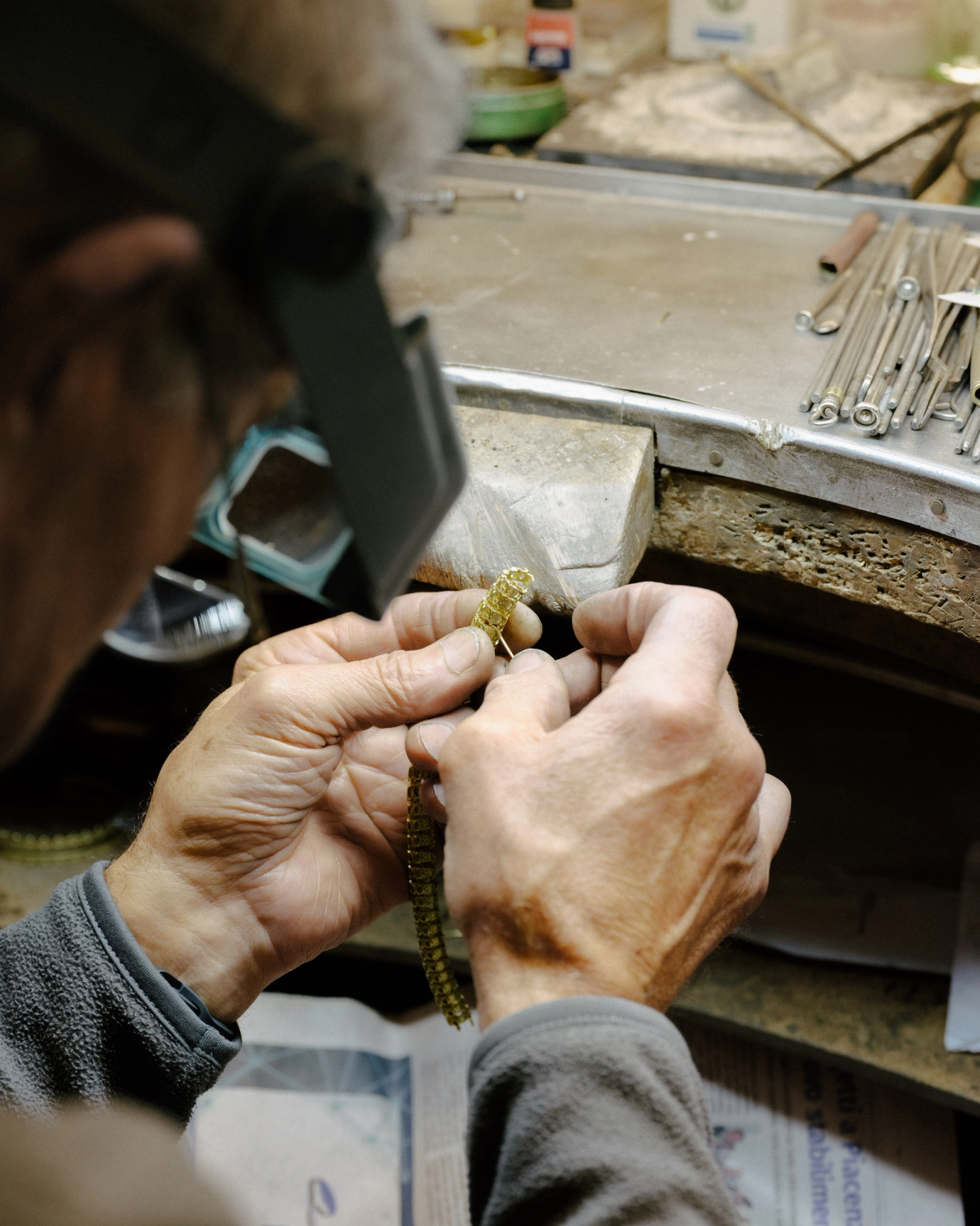 A person wearing a magnifying visor meticulously works on a gold chain at a workbench.