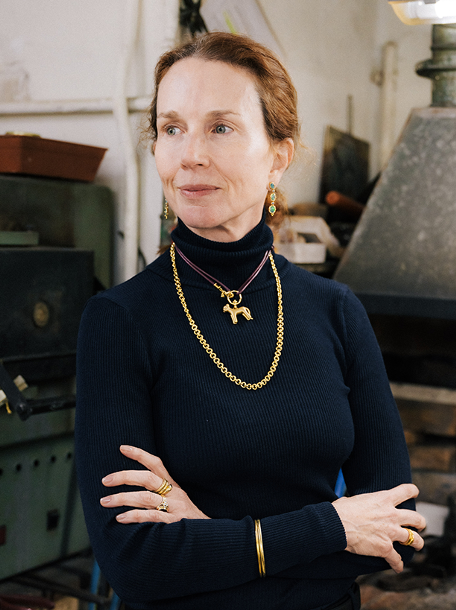 Woman with reddish-brown hair wearing a navy turtleneck, gold dog pendant necklace, and emerald earrings, standing with arms crossed in a workshop.
