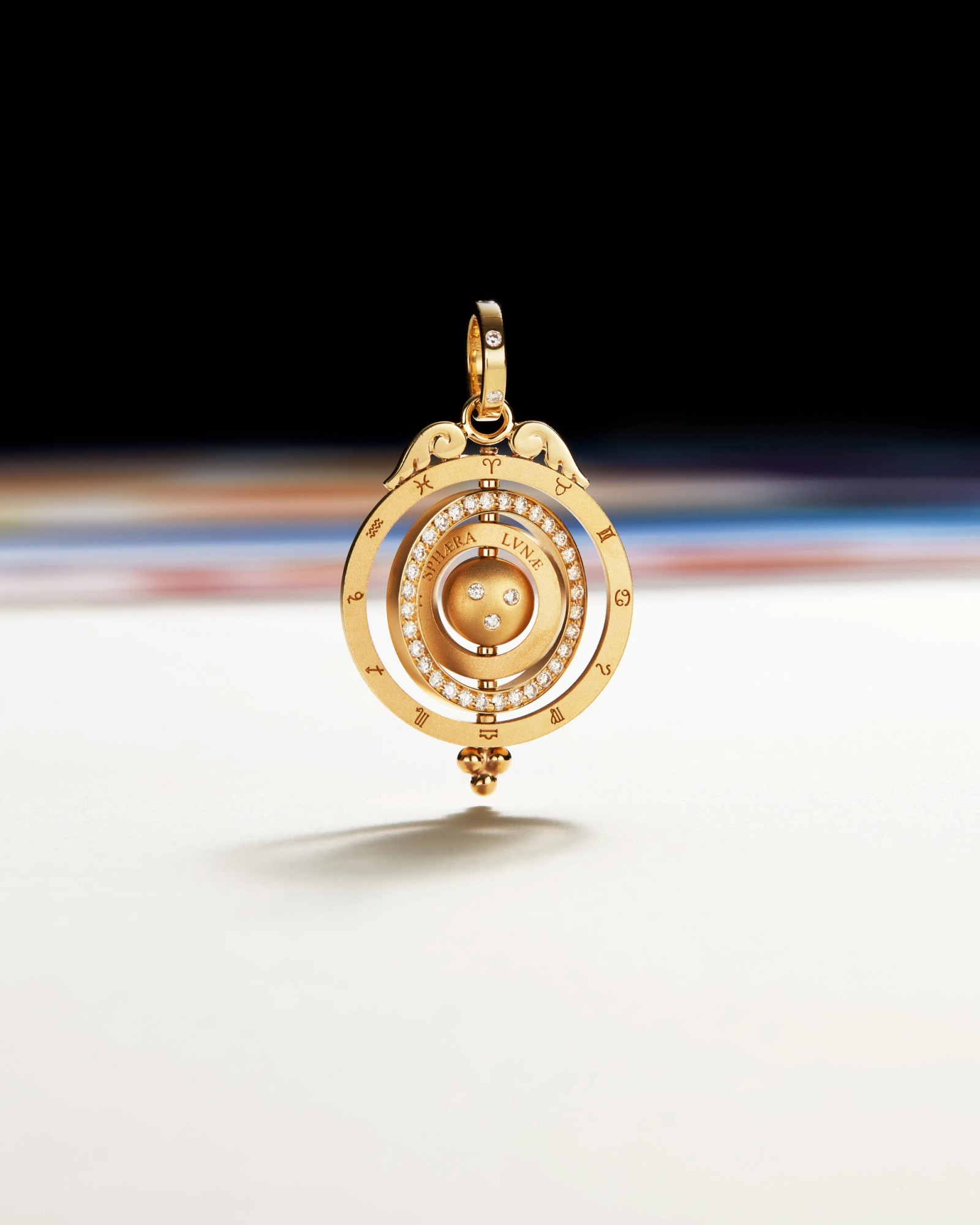 A golden armillary sphere pendant adorned with diamonds and engraved with "SPHERA LVNAE" and zodiac signs, on a white surface with a dark, blurred background.