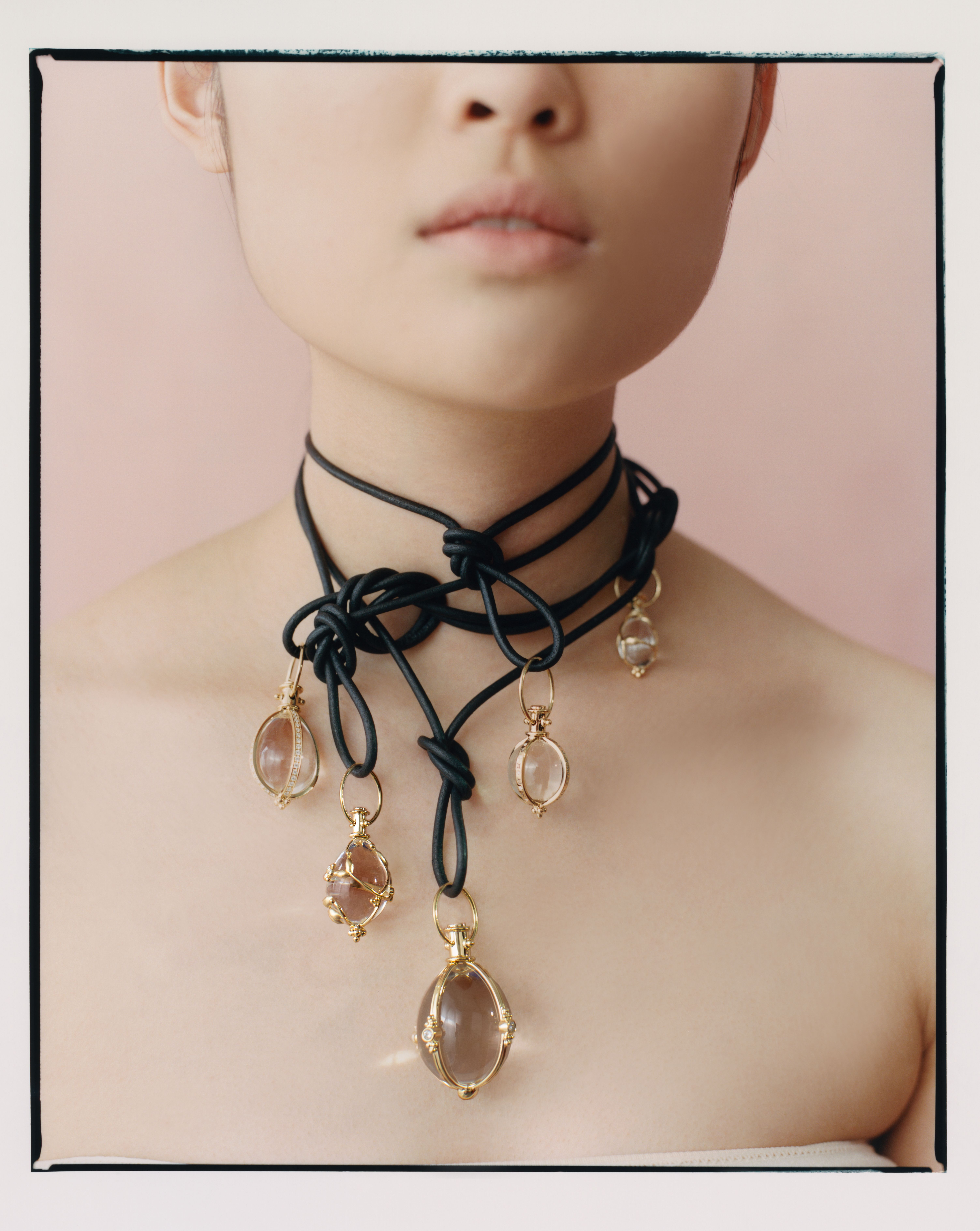 A person wears a black knotted cord necklace with multiple clear stone pendants in gold settings, against a pink background.