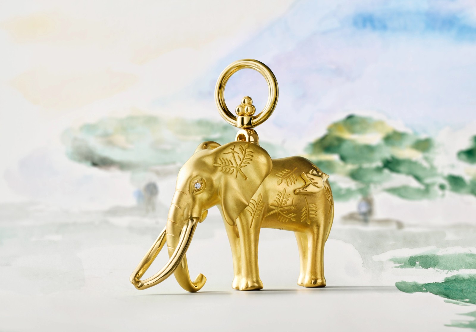 A golden elephant charm with a diamond eye, engraved leaves, and a tiny frog on its back, against a watercolor savanna.