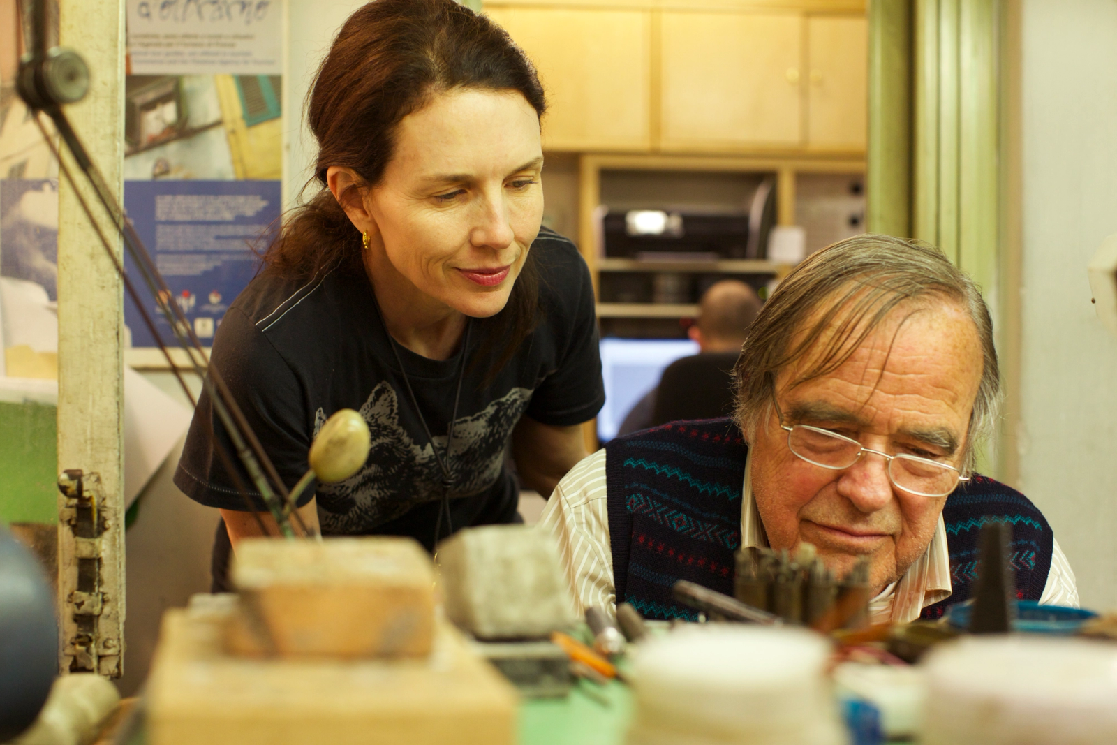 A woman observes an older man working at a workbench.
