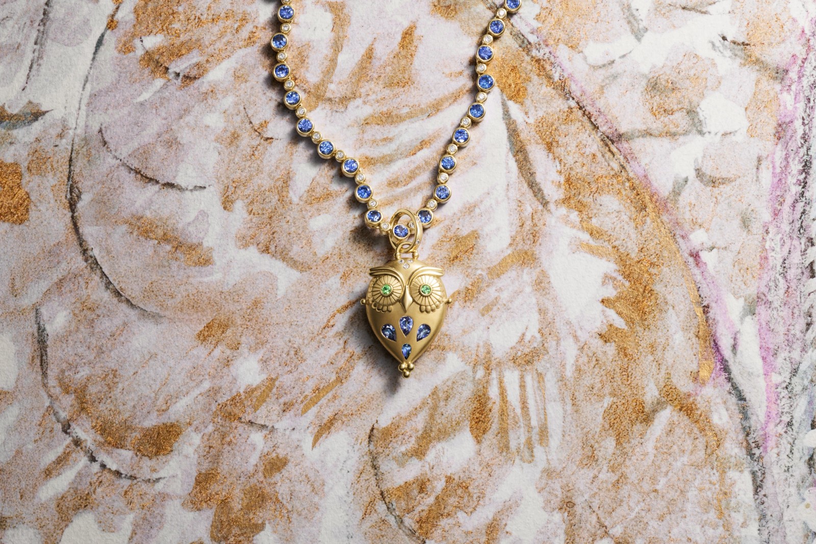 A gold necklace with blue gemstones and a golden owl pendant featuring green eyes, resting on a textured gold and white surface.