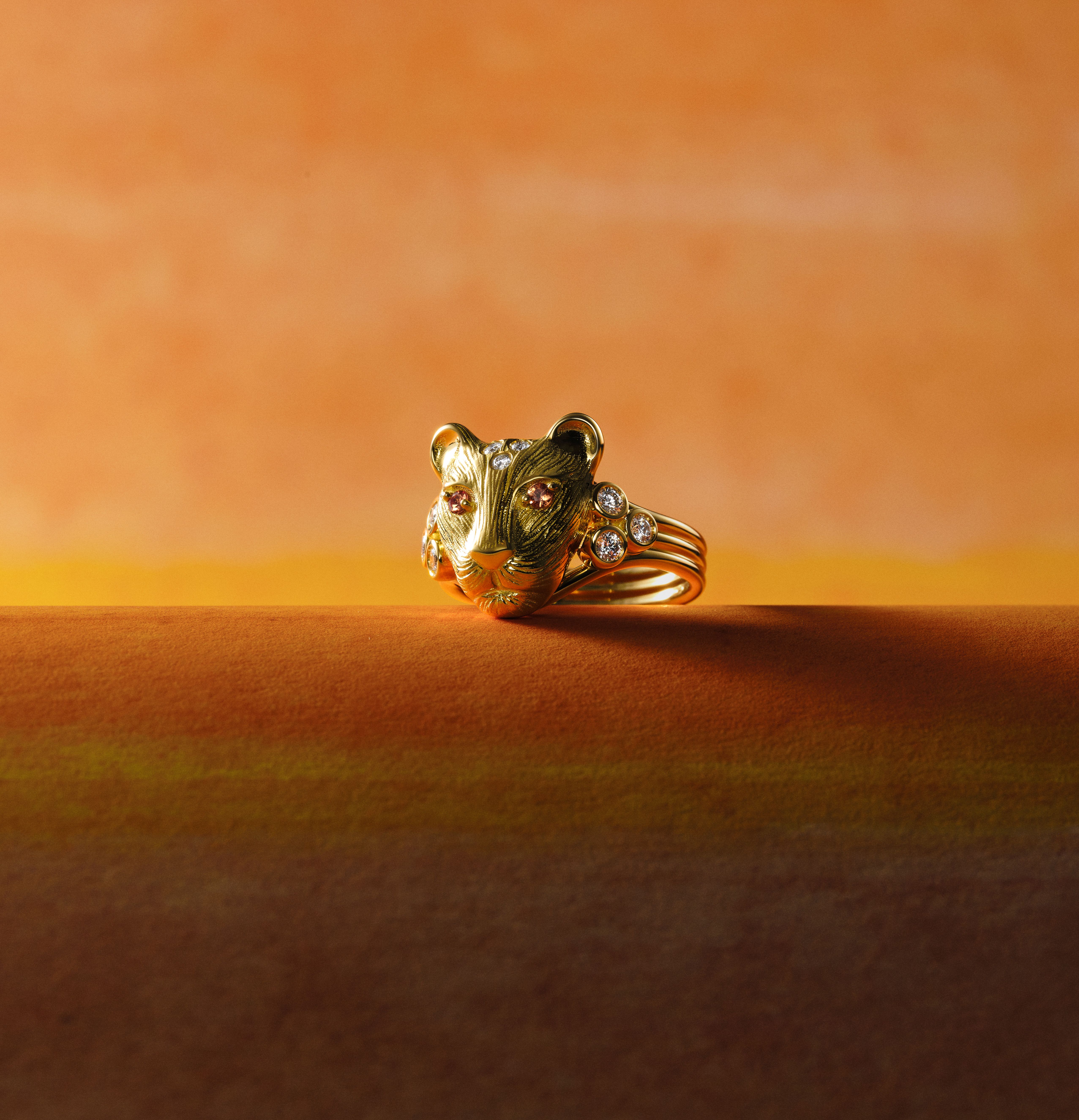 Gold lion head ring with diamonds and pink gemstone eyes on an orange background.