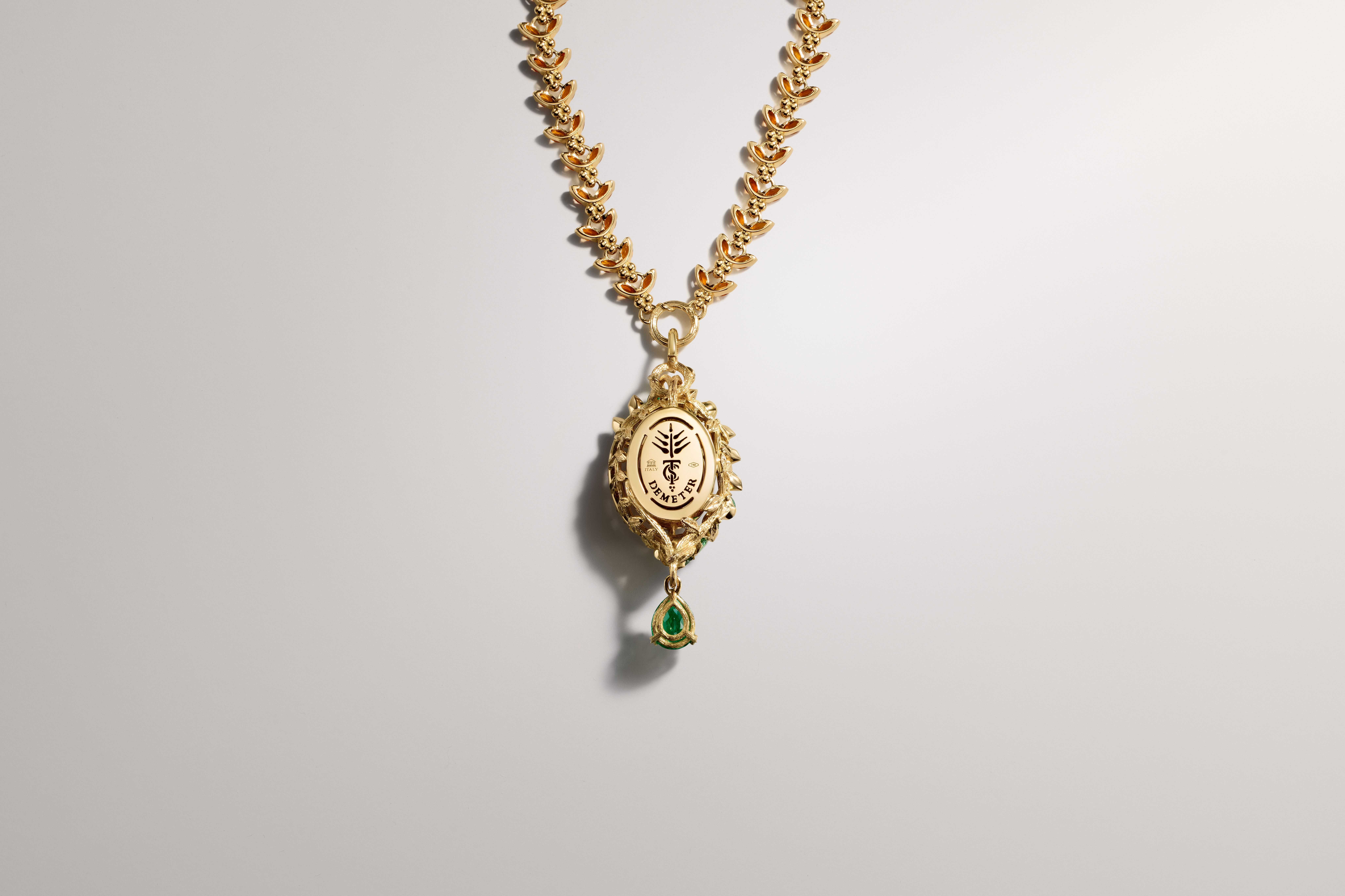 Ornate gold necklace with oval "DEMETER" pendant, emblem, and green teardrop gemstone.