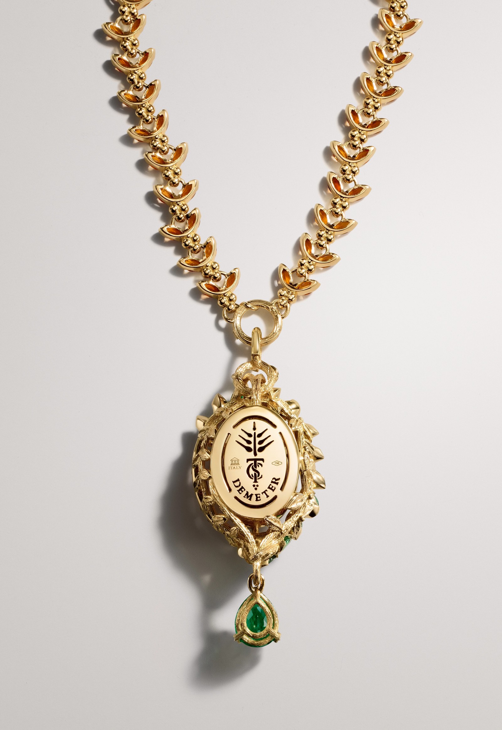 Ornate gold necklace with oval "DEMETER" pendant, emblem, and green teardrop gemstone.