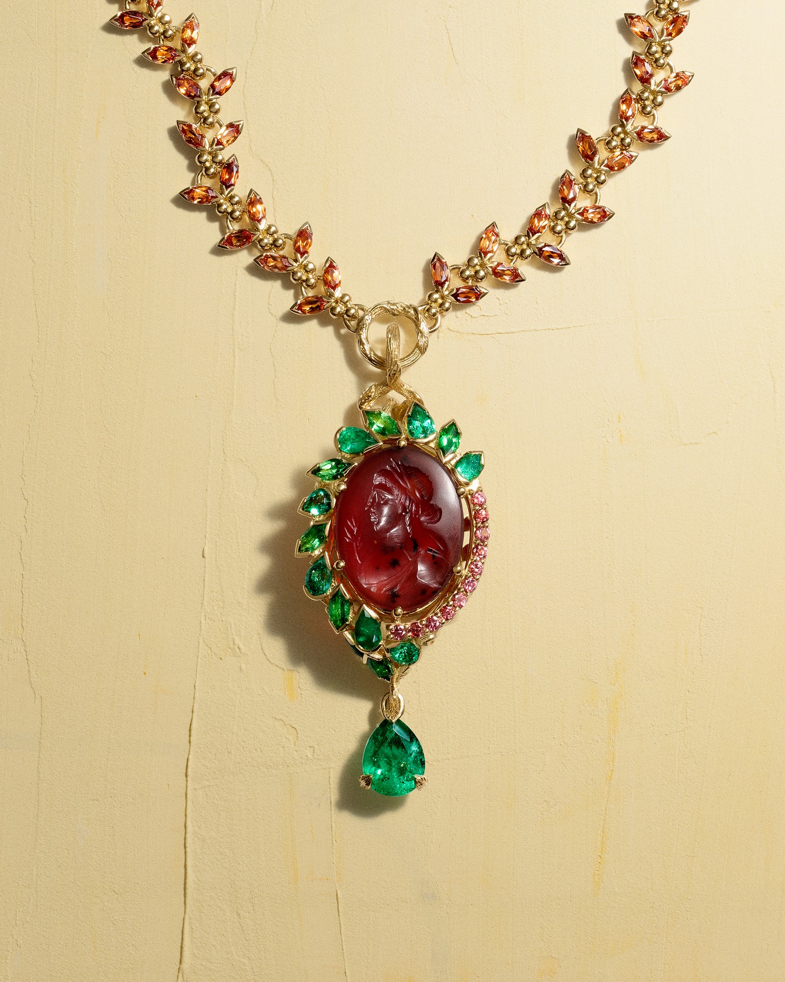 Gold necklace with a chain of orange leaf-shaped stones, featuring an oval red carved cameo pendant framed by green and pink gems, with a dangling emerald.