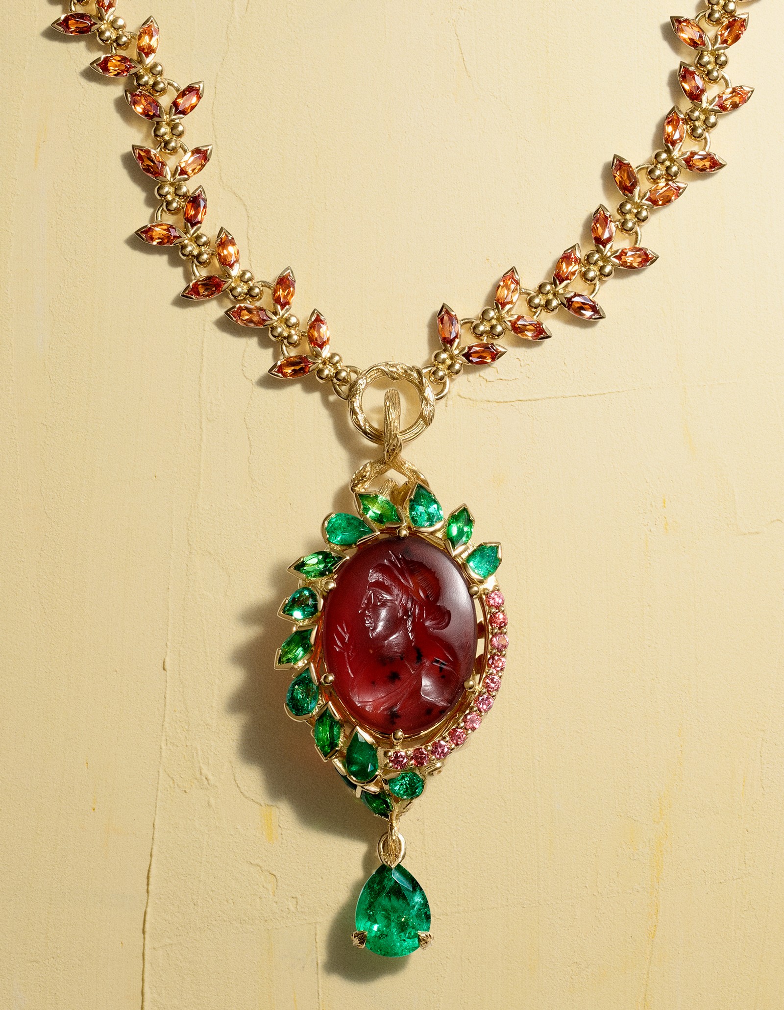 A gold necklace with a leaf-patterned chain set with orange gems, featuring a red intaglio pendant framed by green and pink gems, and a dangling pear-shaped green emerald.