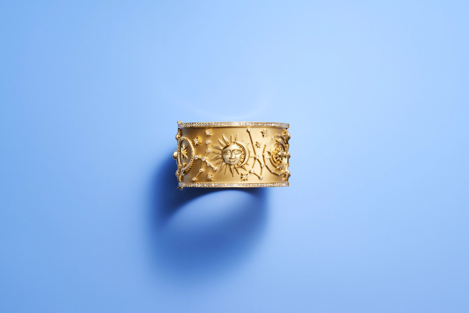 Gold cuff bracelet with sculpted sun, moon, stars, constellations, and diamonds, on a blue background