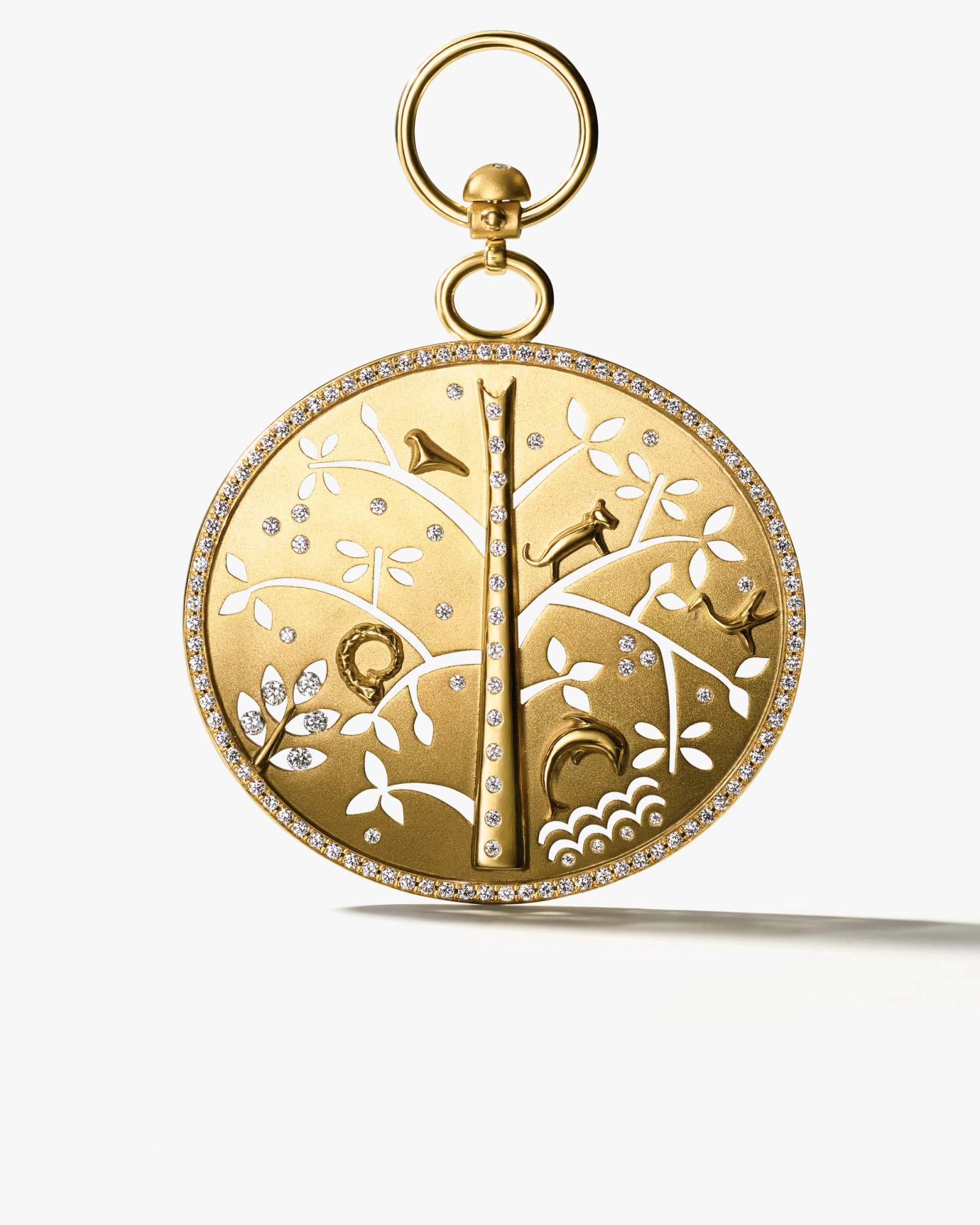 A circular gold pendant with a detailed nature scene, featuring a tree, leaves, a bird, a cat, a snake, and water, with white cut-out details and diamonds.
