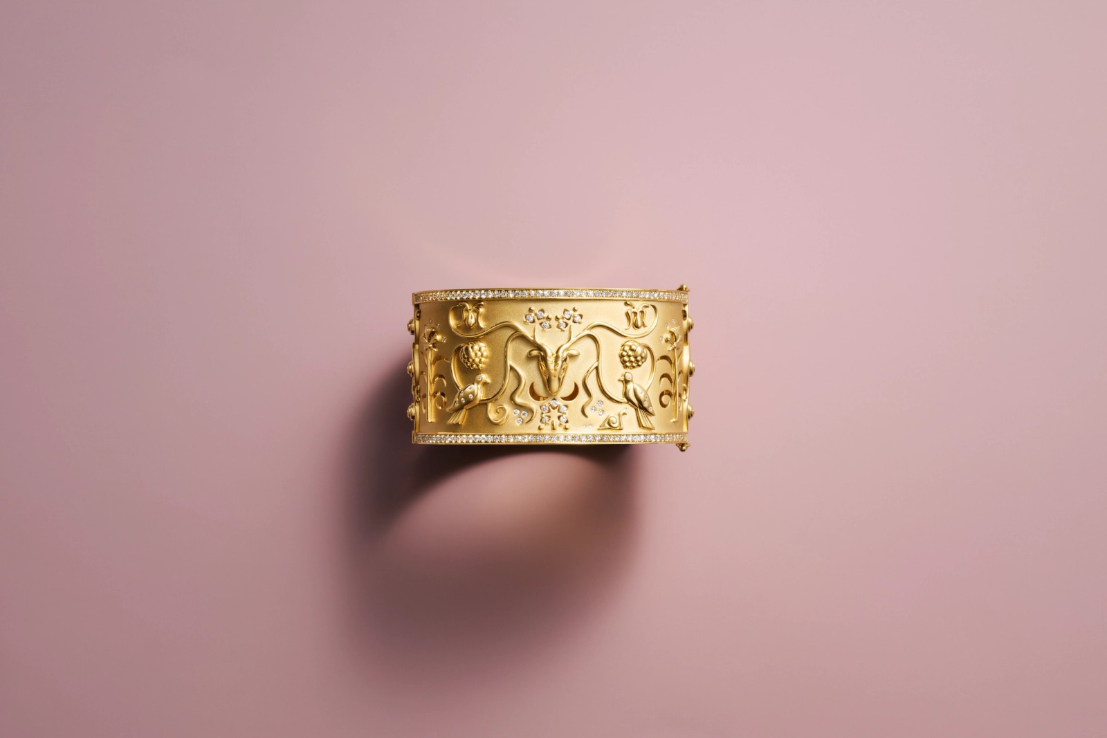 Gold cuff bracelet with embossed botanical design and diamond accents on a pink background.