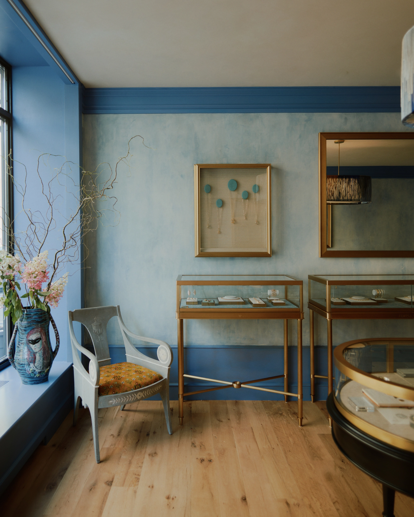 A stylish room with light blue walls, wooden floors, gold-framed glass display cases, an ornate chair, and a vase of flowers.