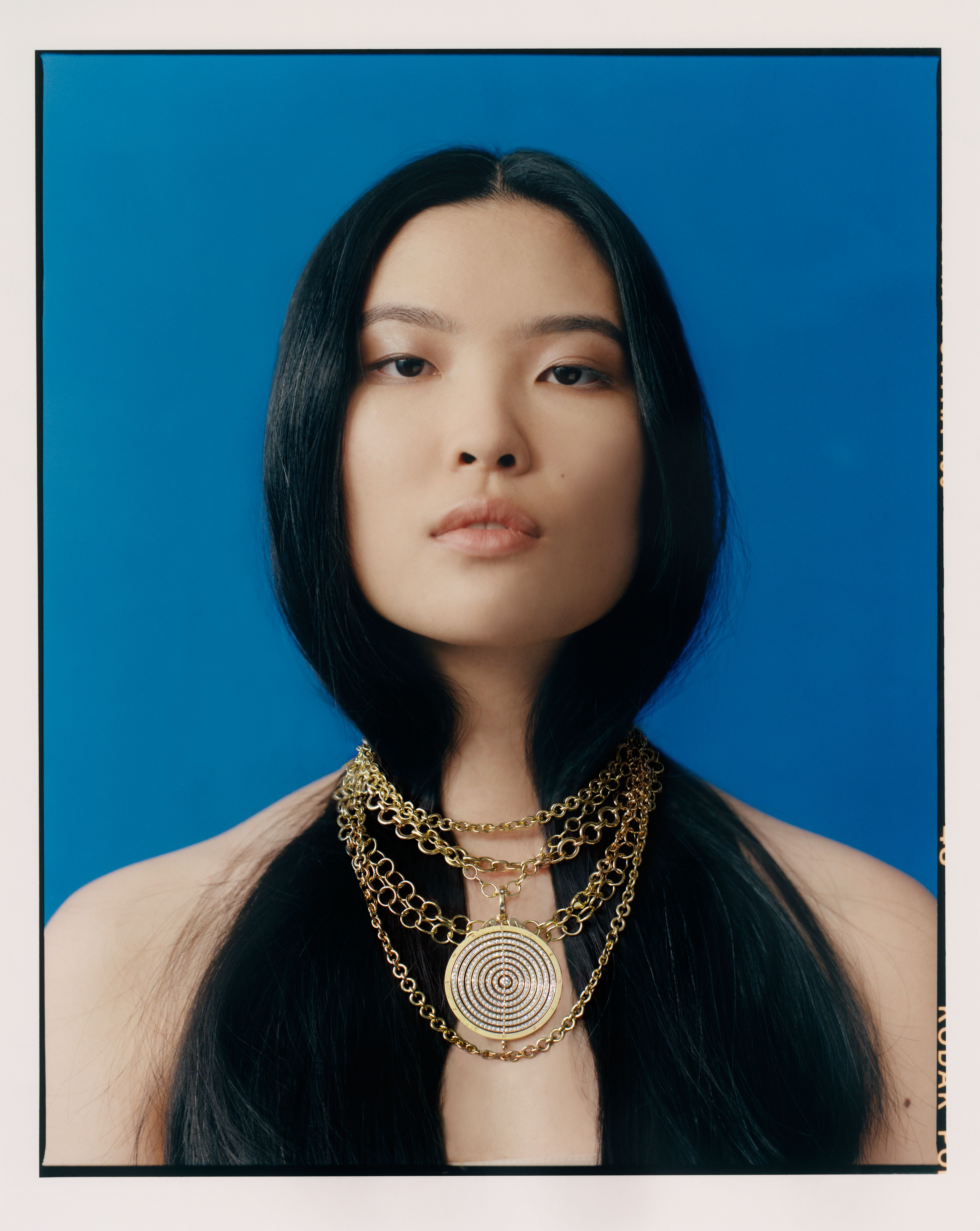 A woman with long black hair, bare shoulders, and a layered gold chain necklace with a prominent circular pendant looks directly at the viewer against a blue background.