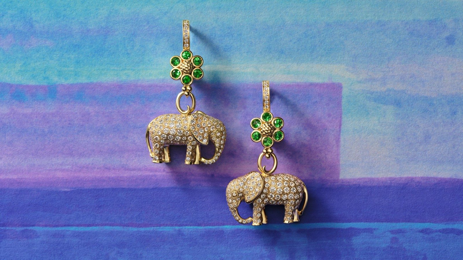 Pair of gold elephant earrings, diamond-encrusted, with green gemstone flower tops on a blue and purple background.