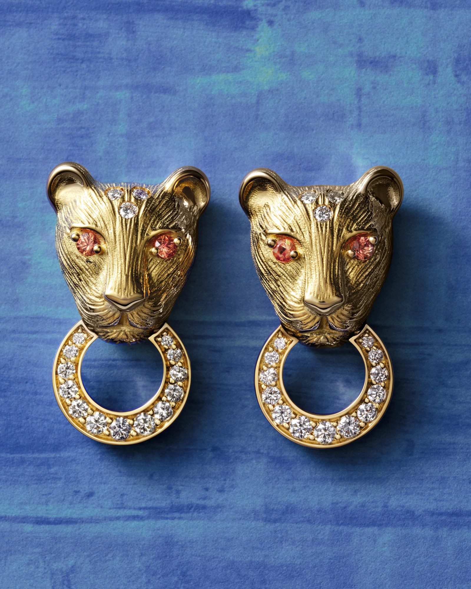 A pair of gold panther head earrings with pink gem eyes and diamond C-hoops, on a blue background.