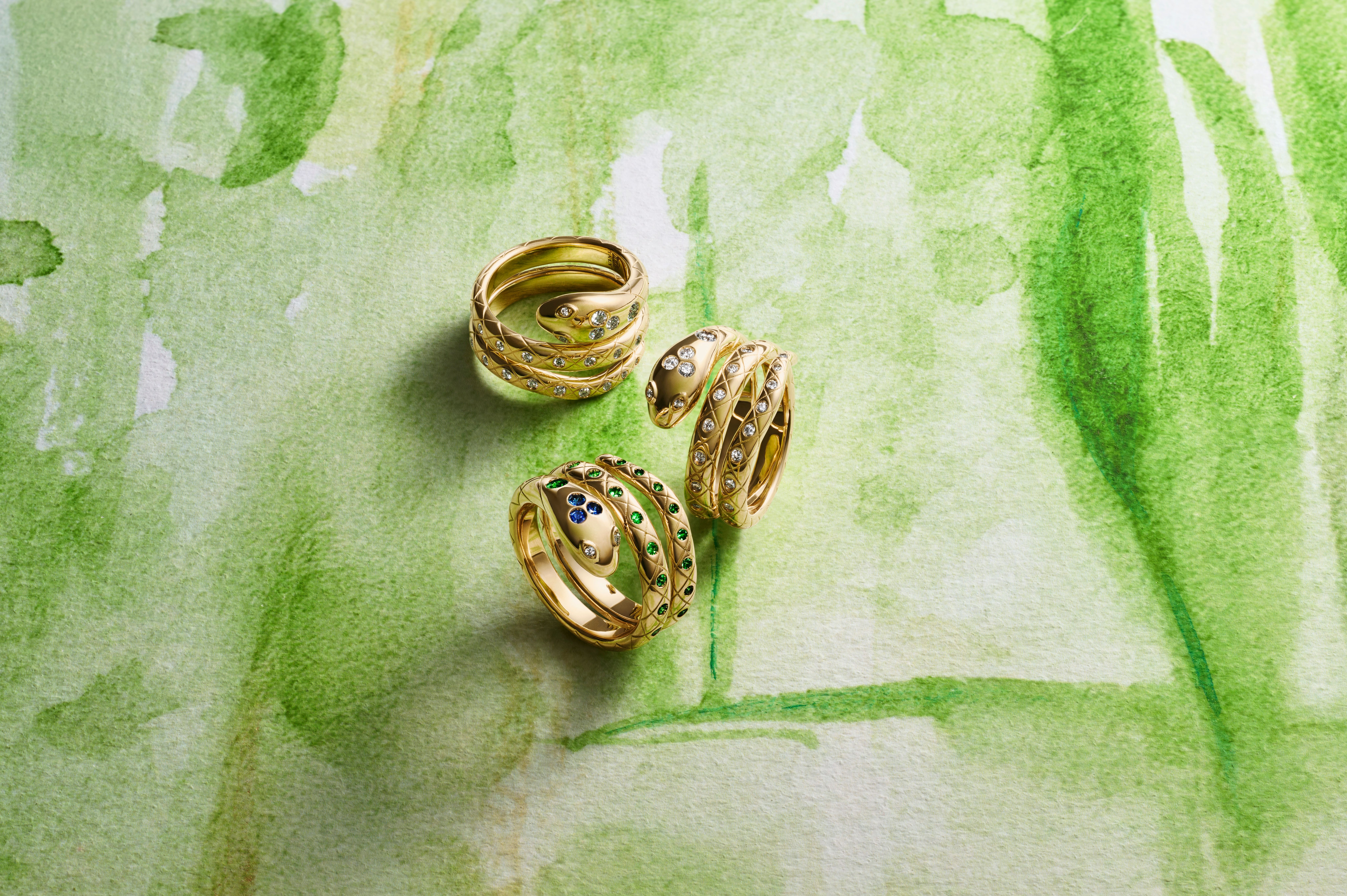 Three gold serpentine rings, two with diamonds and one with sapphires and emeralds, on a green watercolor background.