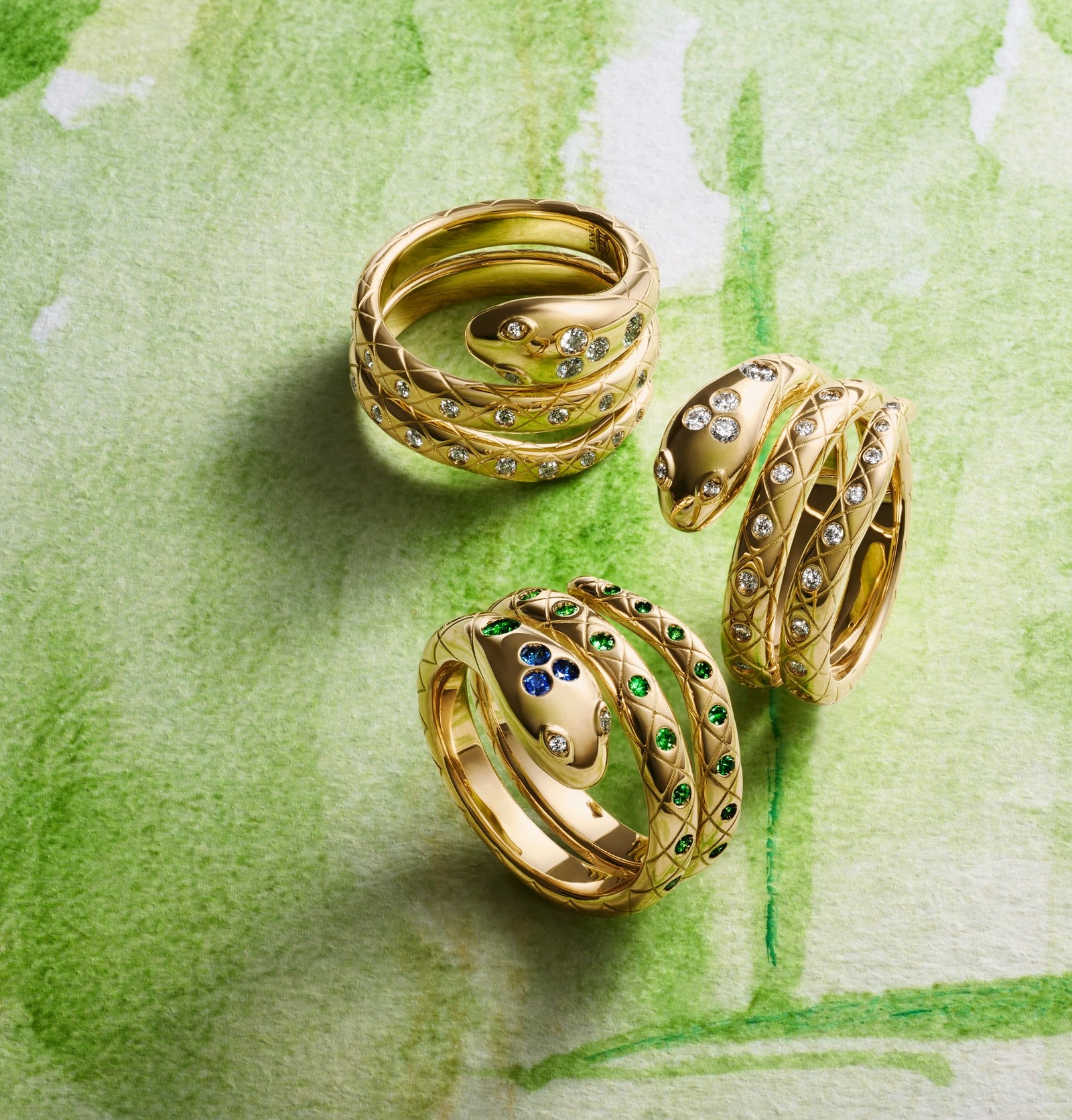 Three gold serpentine rings, two with diamonds and one with sapphires and emeralds, on a green watercolor background.