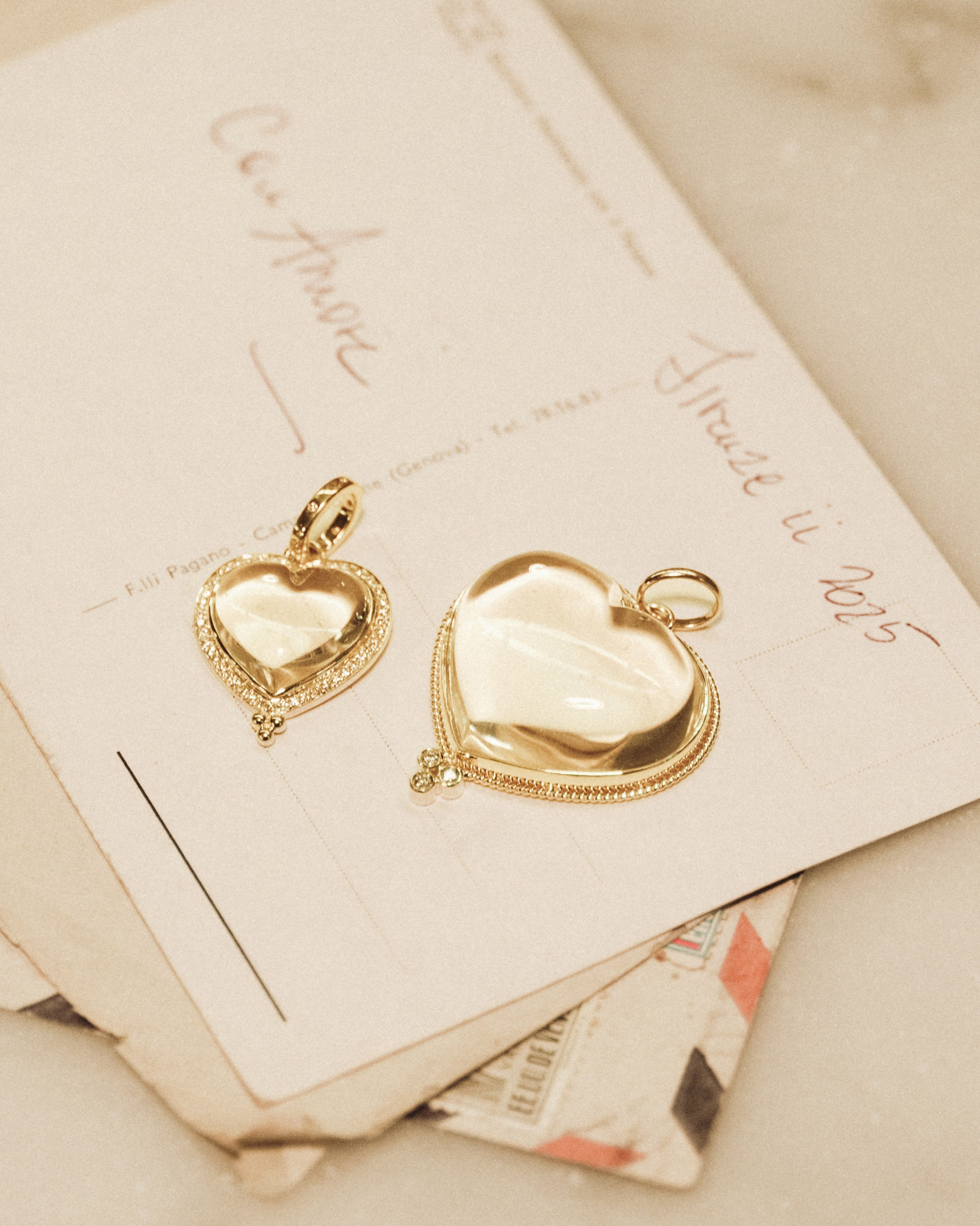 Two golden heart pendants, one with a diamond border, rest on vintage postcards with handwritten Italian text.