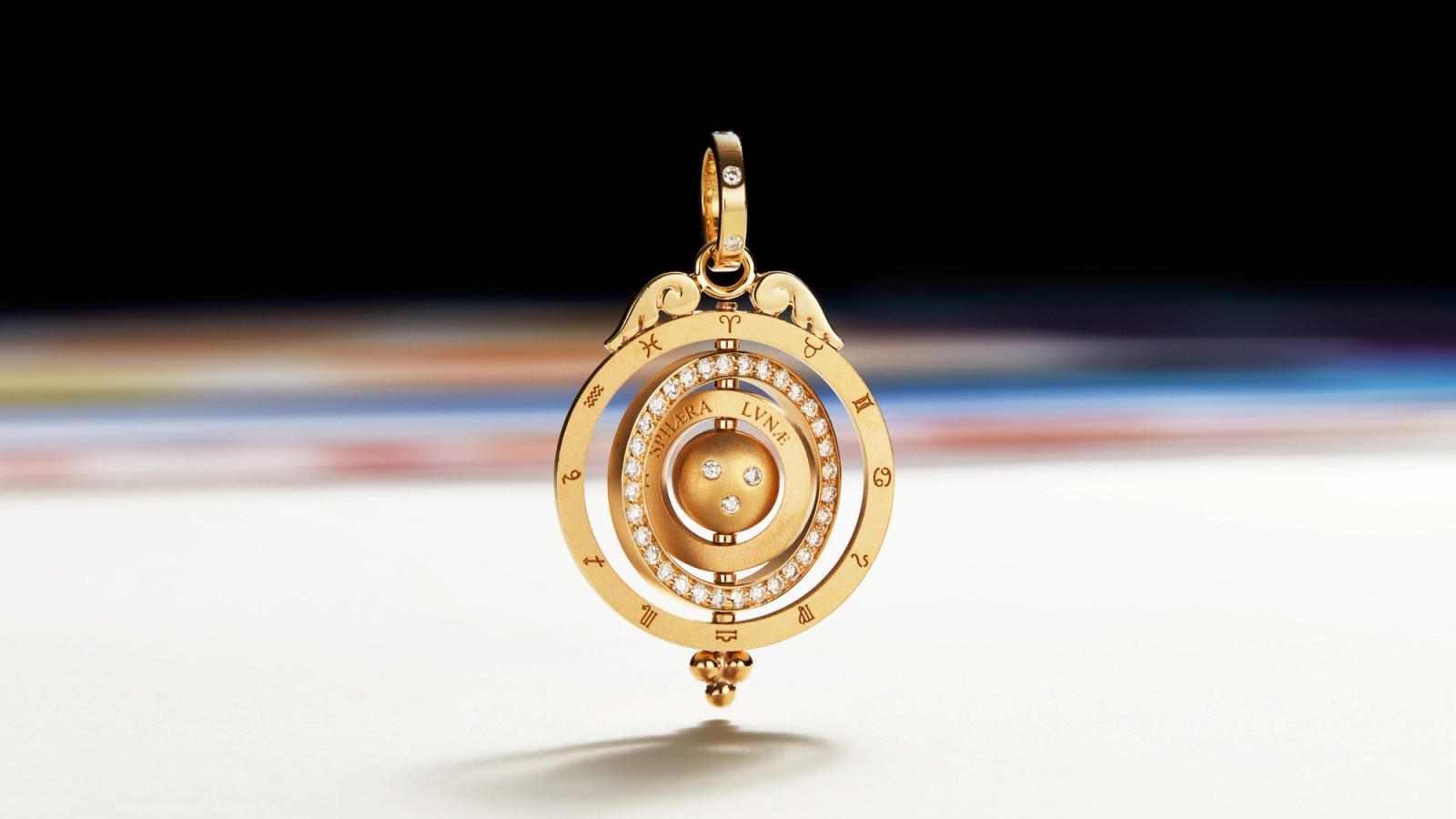 A gold armillary sphere pendant adorned with diamonds and zodiac symbols, against a blurred dark and colorful background.