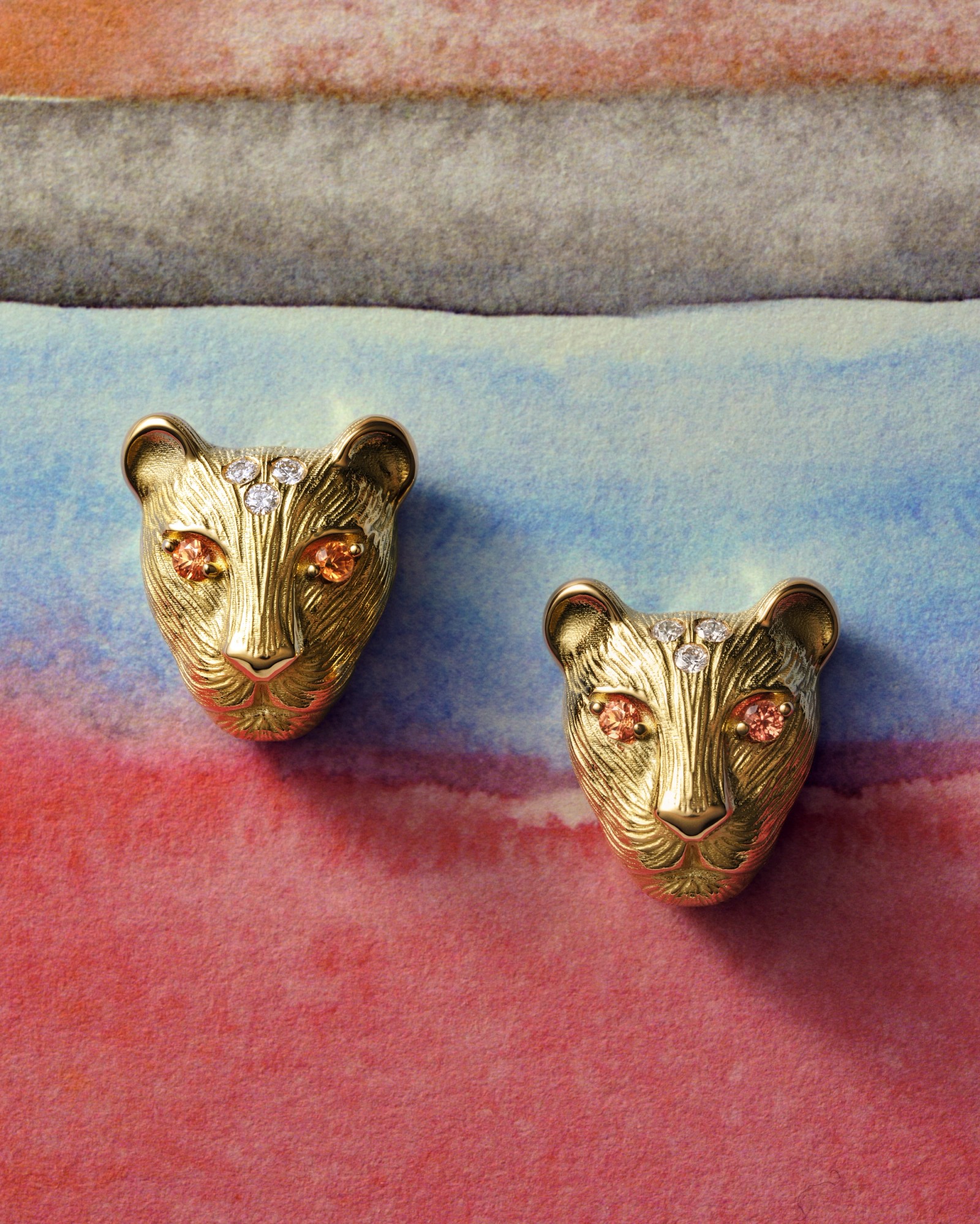 Pair of gold panther head earrings with orange gem eyes and forehead diamonds on a watercolor background.