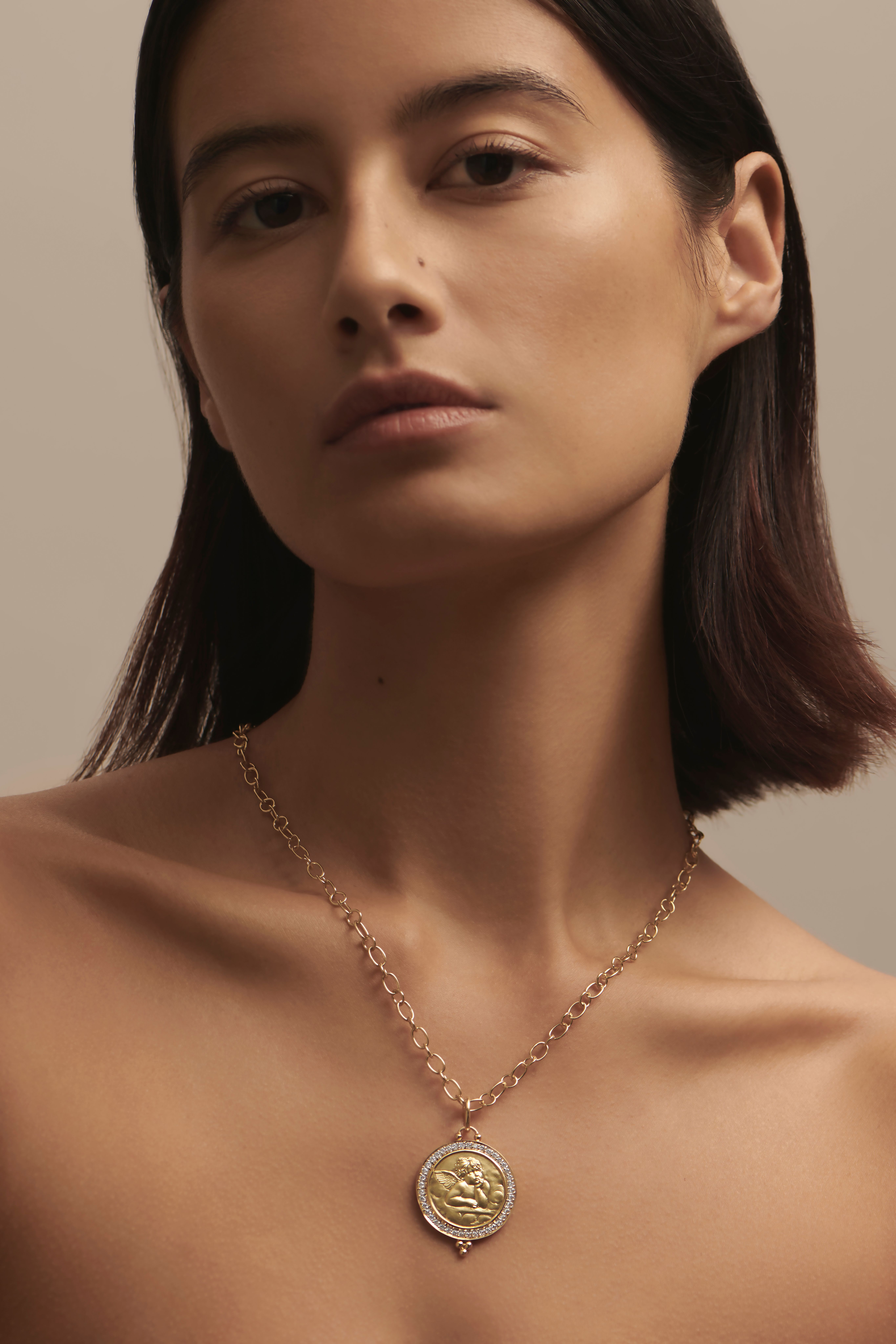 A woman wearing a gold chain necklace with a cherub coin pendant surrounded by diamonds.