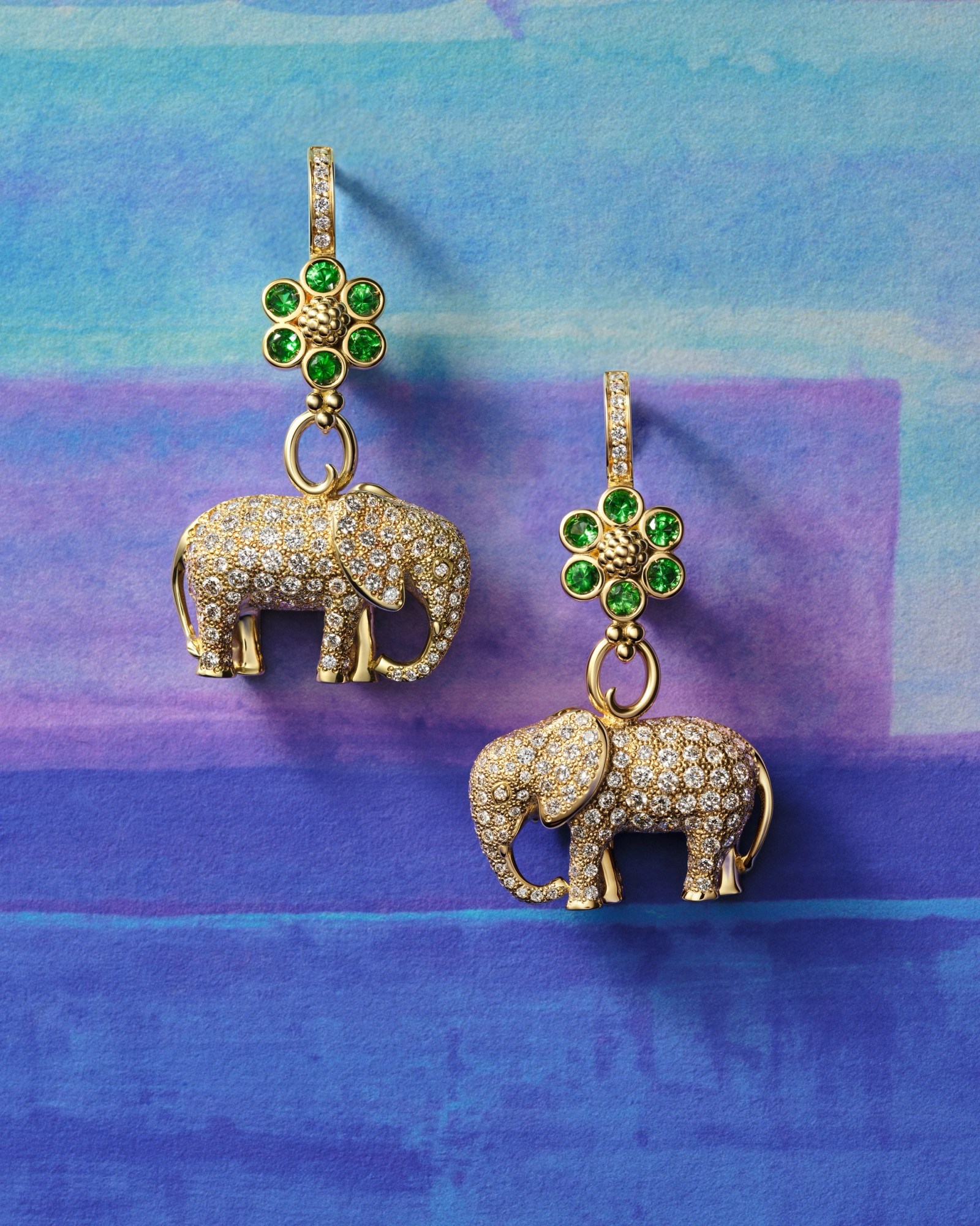 A pair of gold earrings with green gemstone flowers and diamond-encrusted elephant charms on a blue and purple watercolor background.