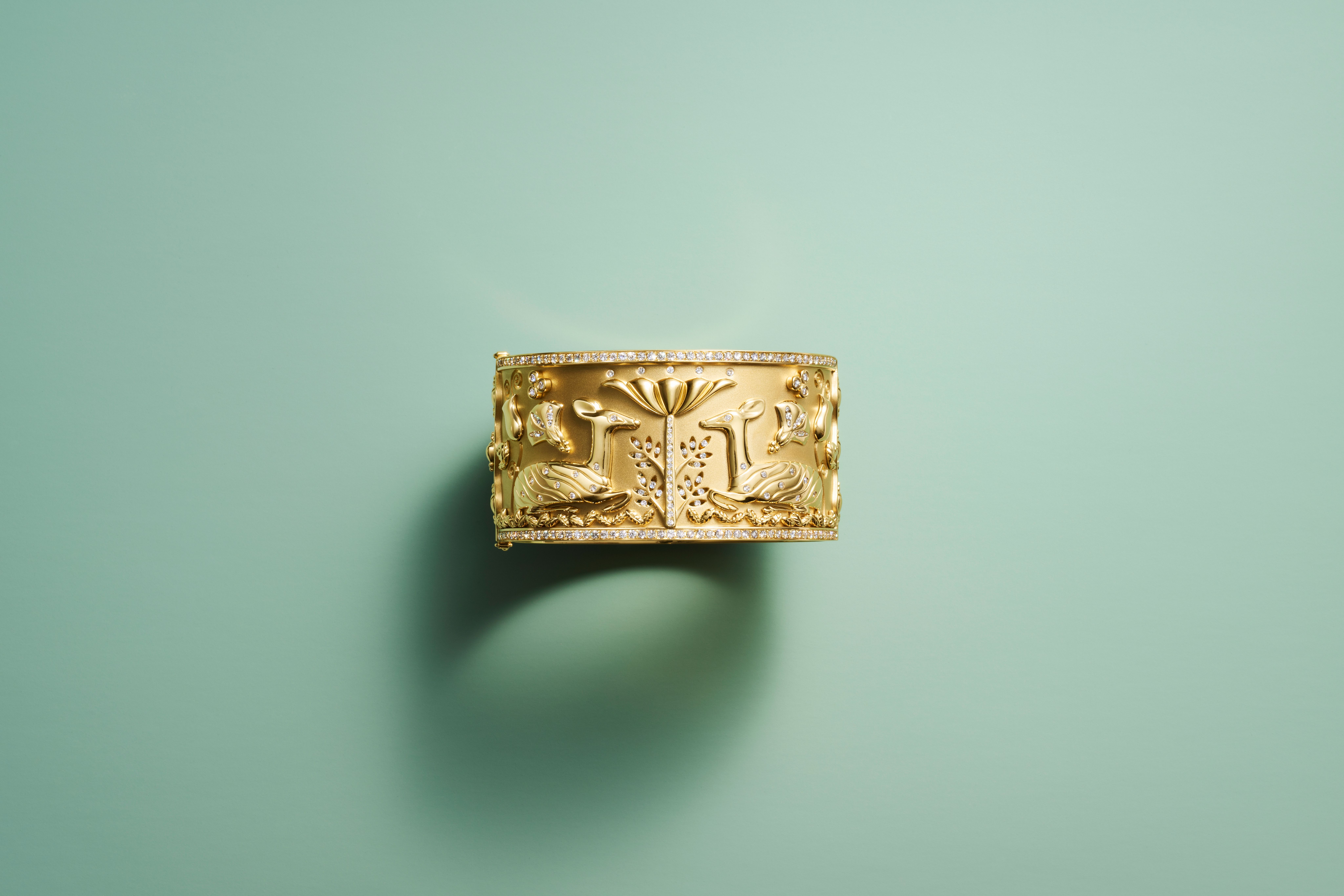 Gold cuff bracelet with raised carvings of lotus flowers, reeds, and a bird, framed by diamond borders, on a mint green surface.