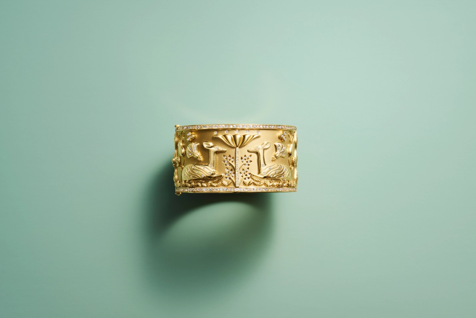 Gold cuff bracelet with carved nature motifs and diamond accents, on a teal background.