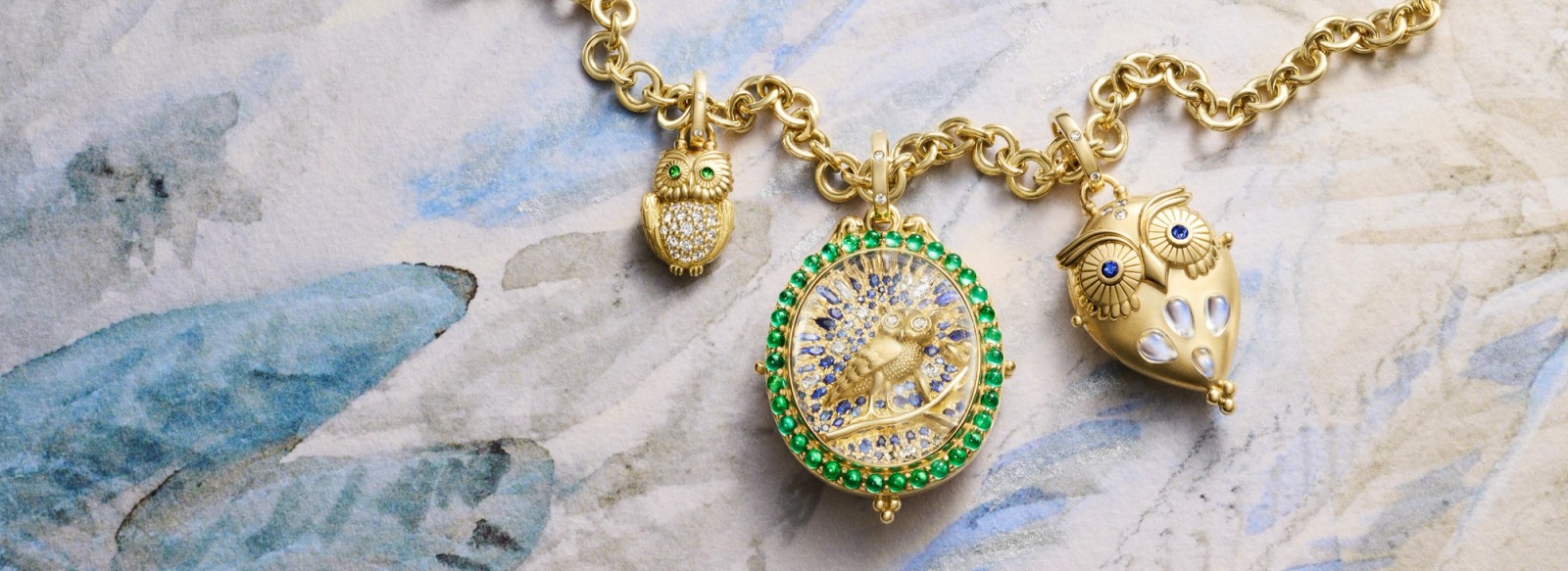 Three gold charms on a chain: a small diamond and emerald owl, a circular locket with a golden bird and blue gems, and a larger gold owl with sapphire eyes, on a watercolor background.