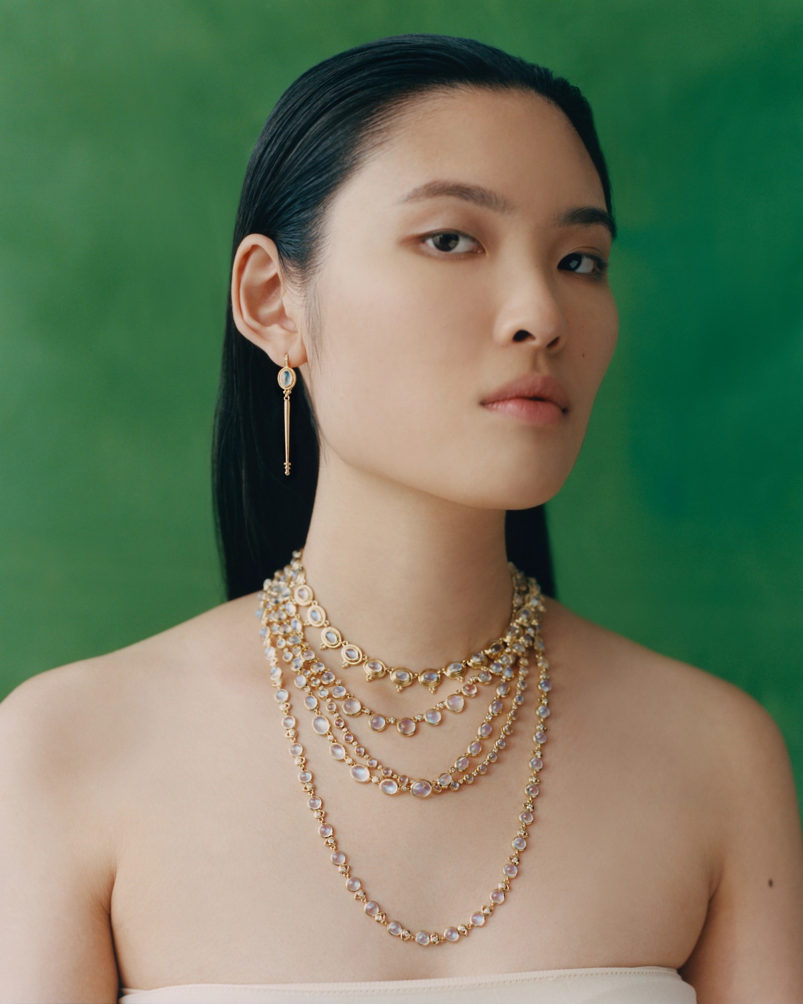 A woman wearing layered gold necklaces with light-colored gems and a matching earring, looking forward against a green background.