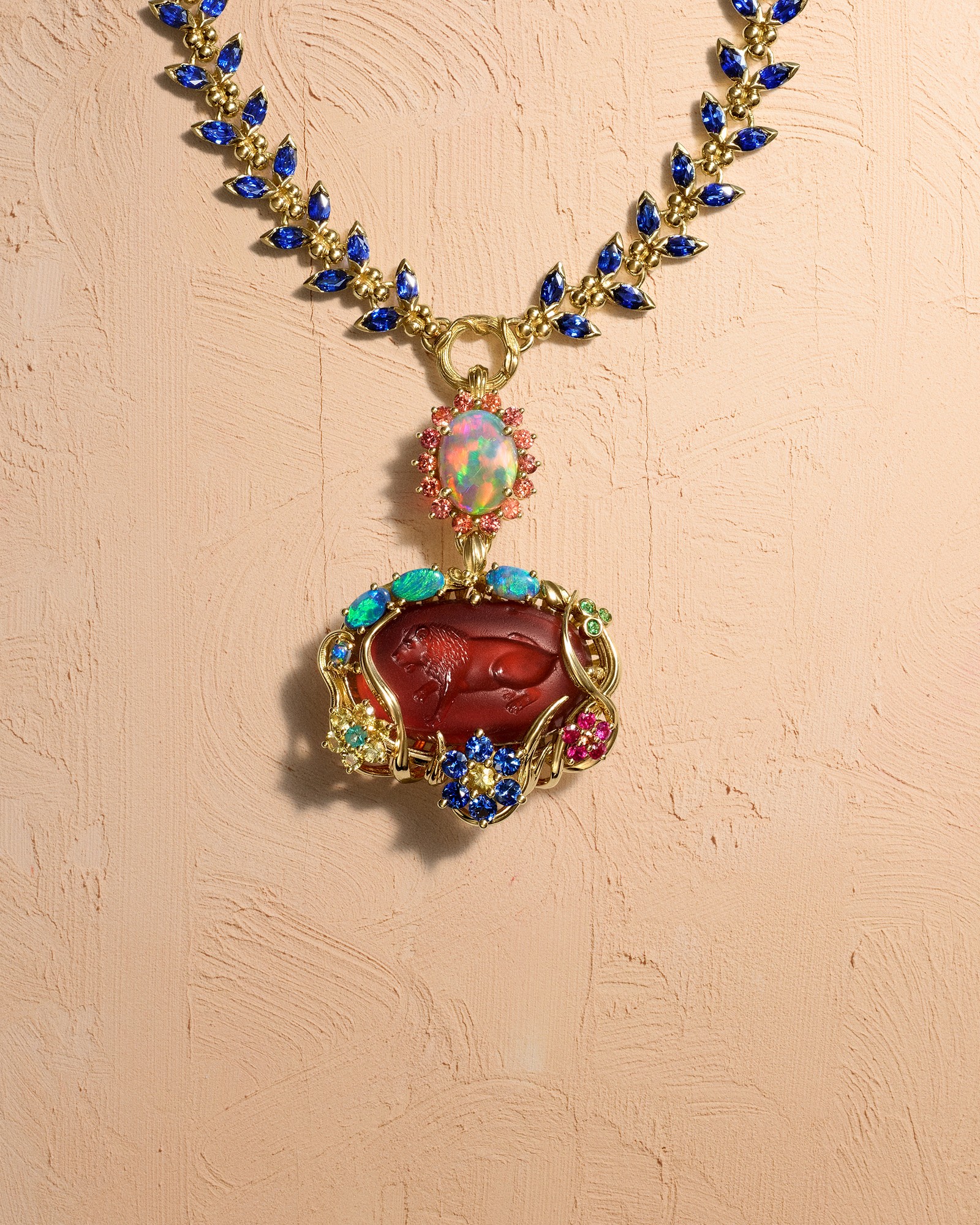 An ornate gold necklace featuring a chain with blue sapphires, a vibrant opal pendant, and a larger red carved gemstone depicting a lion, surrounded by colorful gems.