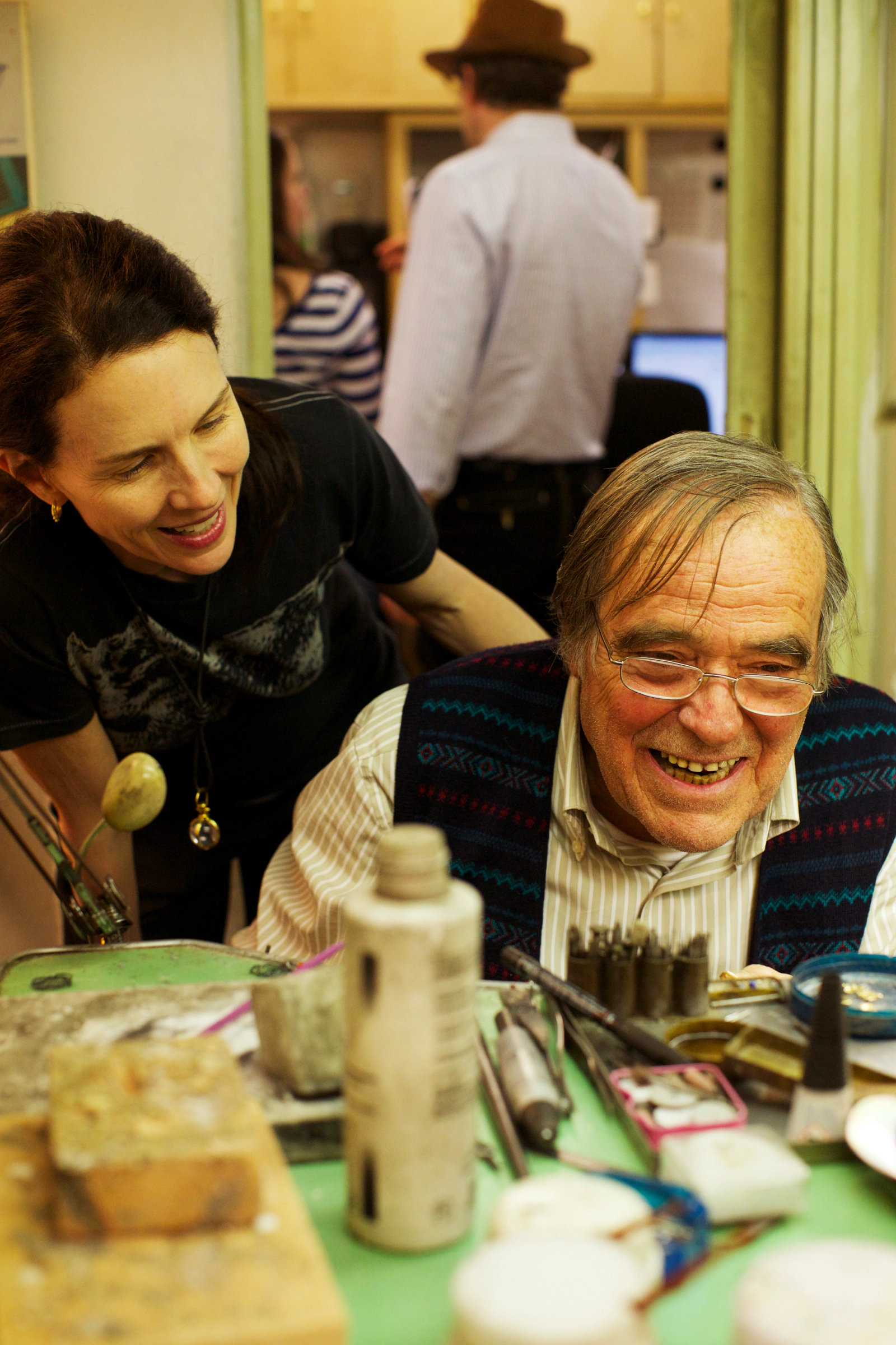 A smiling woman looks at an older man with glasses who is laughing at a cluttered workbench.