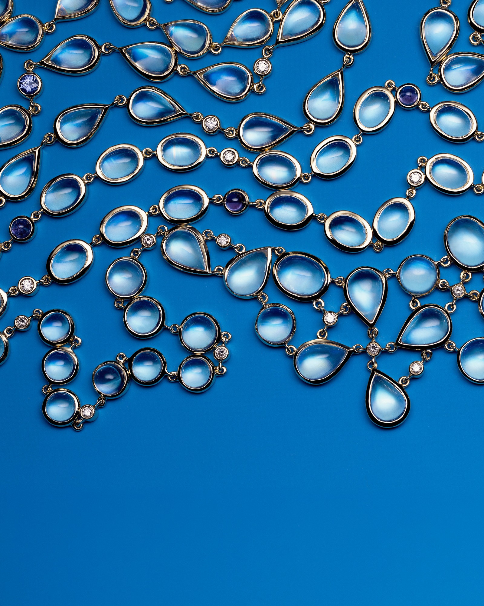 Gold jewelry with light blue oval and teardrop cabochon gemstones, diamonds, and purple stones, laid on a blue surface.